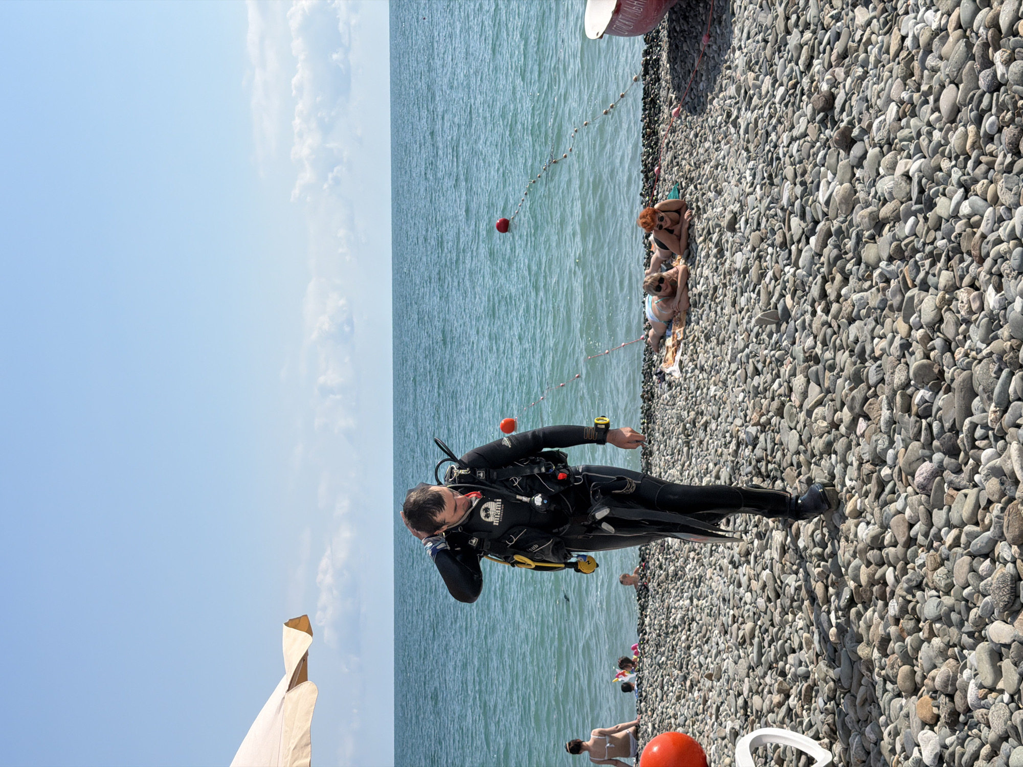 A male diver in a full black wetsuit with yellow accents walks across a pebble beach towards the camera. He is touching his head with his right hand. The diver is equipped with a scuba tank and regulator. In the background, the turquoise sea is calm under a clear blue sky with scattered clouds. A string of buoys floats on the water. Several people are visible on the beach and in the shallow water. Two women are sunbathing on towels on the beach, and a woman in a white swimsuit is sitting further back. Red buoys are present on the beach and in the water. A portion of a red boat is visible on the right side of the frame. The scene is set at a beach in the city of Sirius, Russia.
