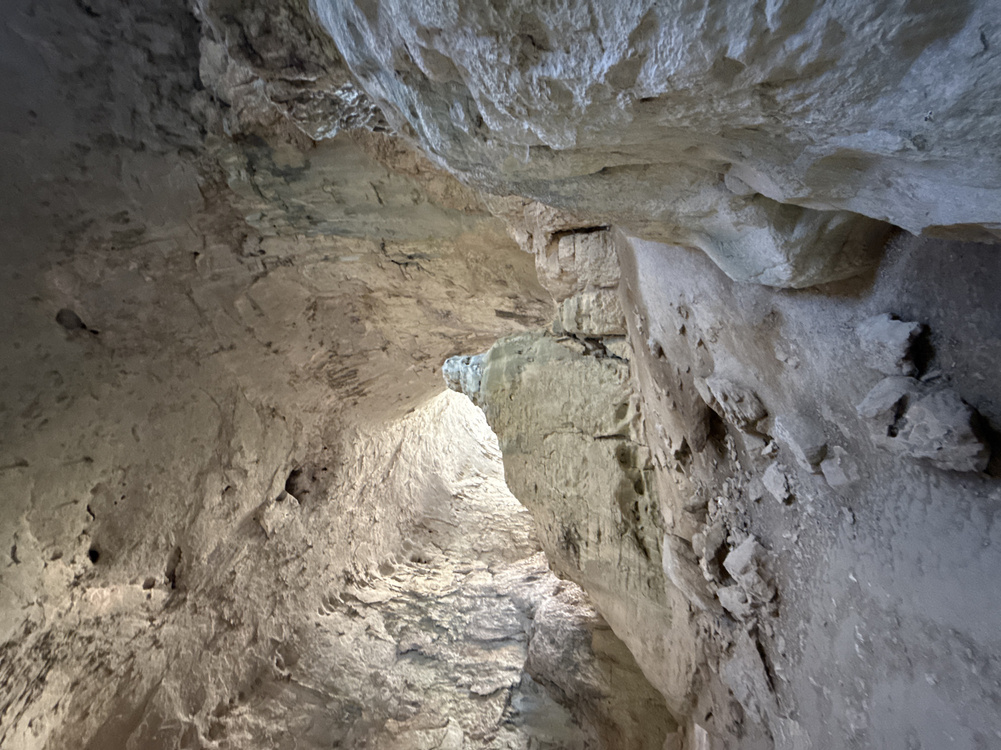The image shows the interior of a cave, likely the Karlukovo Caves in Bulgaria. The cave features a large, cavernous space with rough, textured rock formations. The ceiling is high and arched, with visible cracks and irregularities in the rock. Natural light illuminates the cave from the far end, creating bright highlights and shadows on the surfaces. The floor of the cave is covered in dirt and loose rocks, with a path winding through the formations. Several large rock pillars and boulders are present, some appearing to be stalagmites or other mineral deposits. The overall impression is one of a vast, ancient underground space.