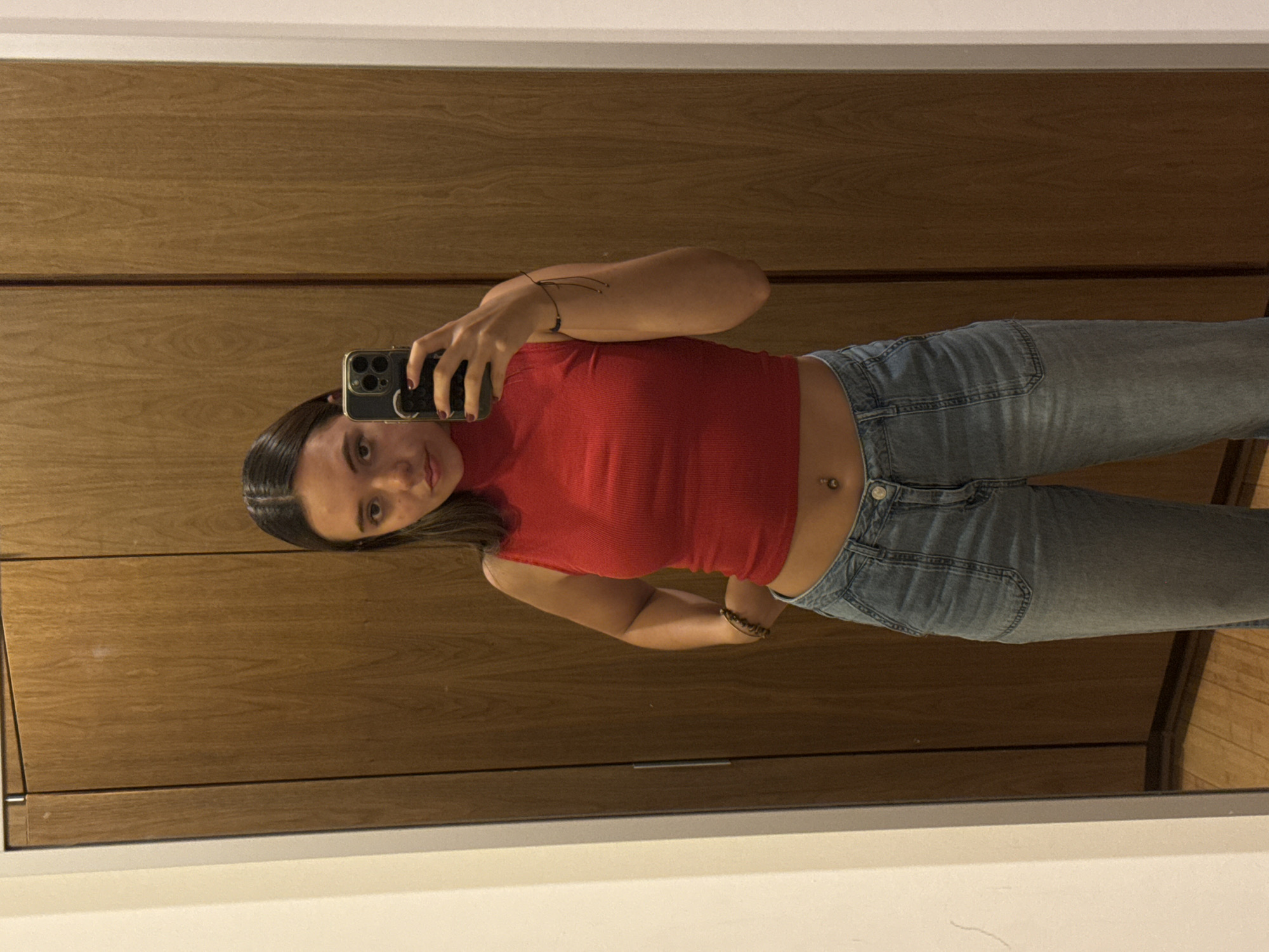 A young woman stands in front of a mirror and takes a selfie. She is wearing a red crop top and light blue jeans. Her phone is an iPhone. The background shows a light-colored wood panel, suggesting an interior space, possibly a bedroom or dressing area. The location is San Bernardino Tlaxcalancingo, Mexico.