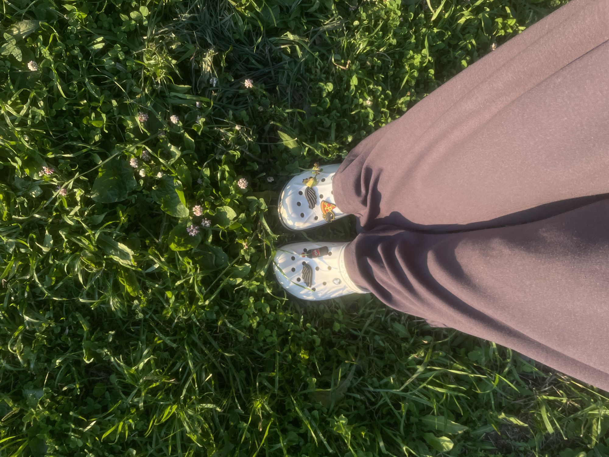 The image shows a person's legs and feet, wearing white Crocs with charms, standing on a patch of green grass. The Crocs have charms including a slice of pizza and a Coca-Cola bottle. The person is wearing dark purple sweatpants. The grass is lush and green, with some small pink clover flowers interspersed. The scene is illuminated by sunlight, creating shadows and highlighting the texture of the grass and fabric. The image is taken from a top-down perspective. The location is identified as Podshivalovo, Russia.