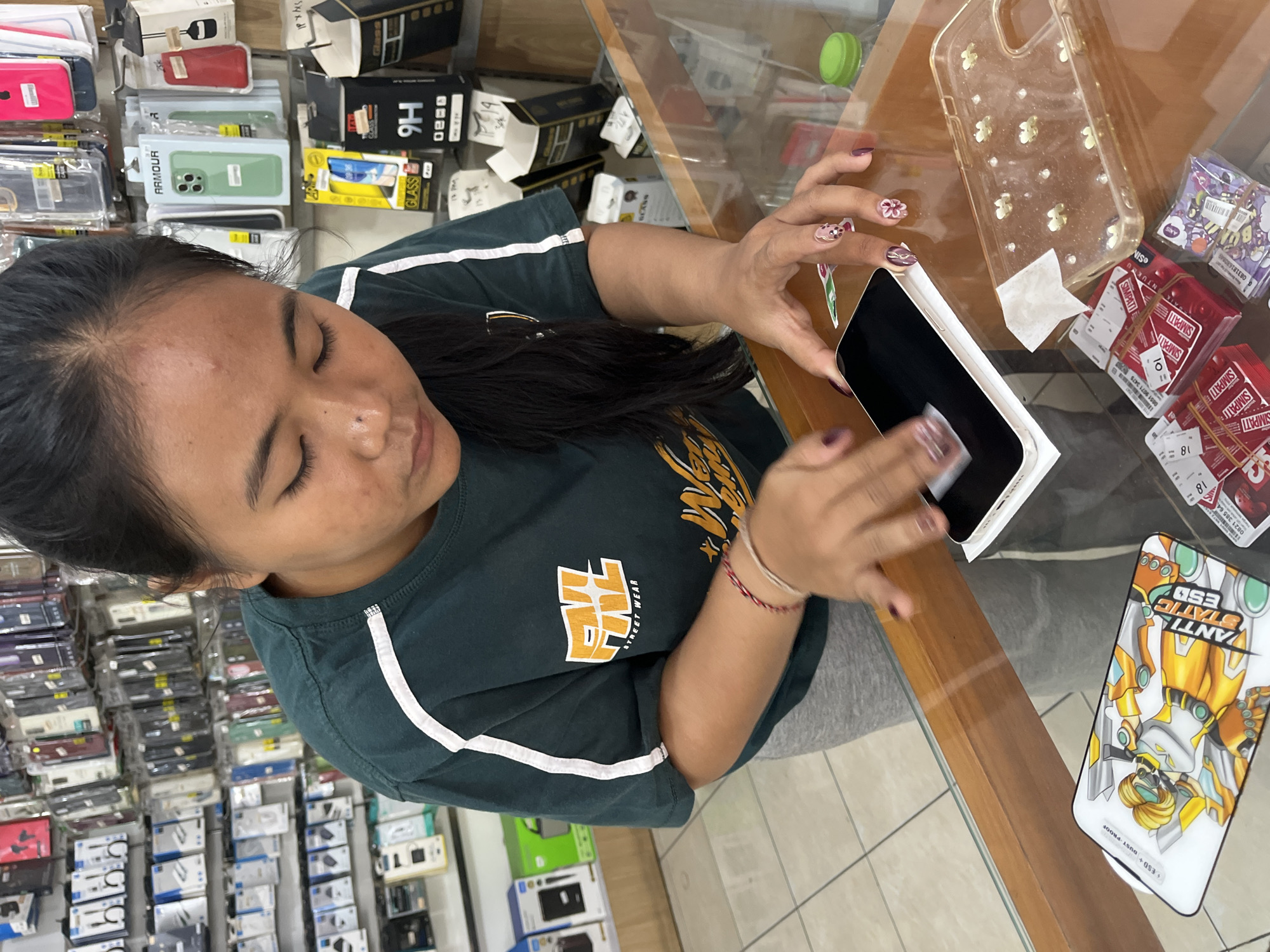 In the city of North Kuta, Indonesia, a young woman with dark hair, wearing a dark green t-shirt with white stripes on the sleeves, is intently cleaning a white smartphone with a small white cloth. Her fingernails are painted with a pinkish-purple polish and adorned with small flower designs. On the glass counter in front of her, to her right, is a clear phone case filled with small white flower embellishments, and to her left, several packs of red SIM cards are neatly arranged. Behind her, shelves are packed with various electronic accessories, including phone cases, chargers, and screen protectors. The environment appears to be an electronics store.