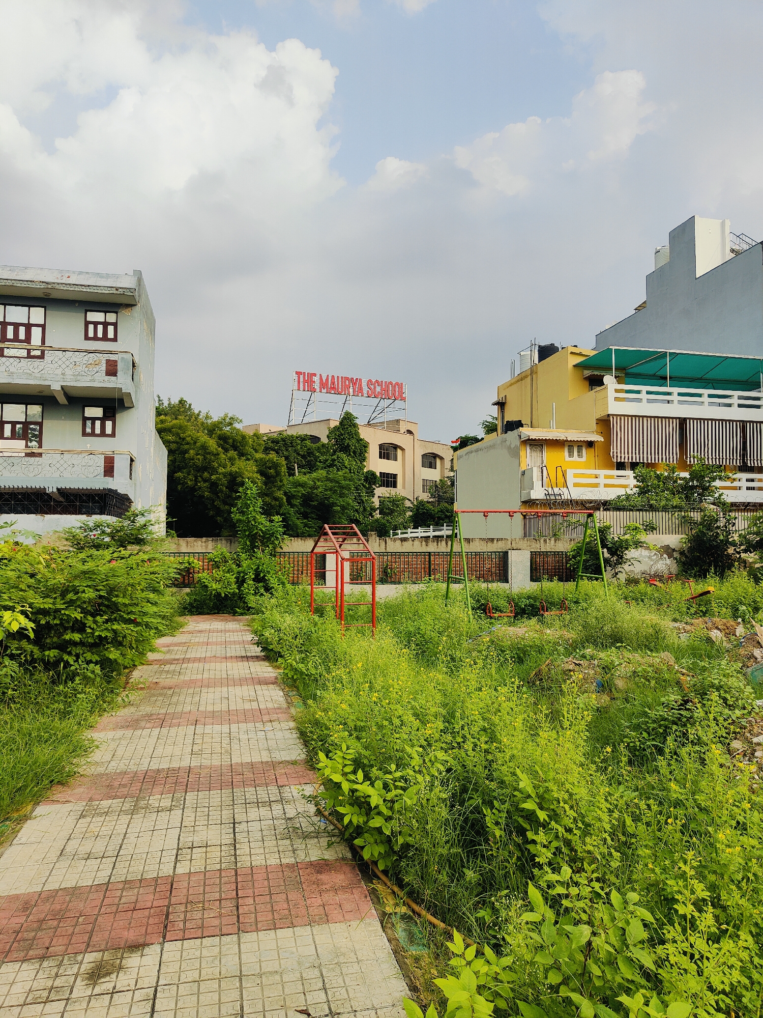 The image captures a scene in Gurgaon, India, featuring a pathway leading into an overgrown area with playground equipment. On the left, a multi-story building with a light blue facade and red-brown window frames stands. Ahead, a red sign with white letters spells out "THE MAURYA SCHOOL." Behind the school sign, various buildings are visible, including a yellow one with striped awnings. In the foreground, a checkered pathway of red and white tiles winds through lush green vegetation. To the right of the pathway, a red metal frame, possibly part of a climbing structure, and a green swing set with two red swings are present. The ground is covered in tall, wild grass and other plants, suggesting a somewhat unkempt or natural state. The sky is partly cloudy, with white clouds against a light blue background.