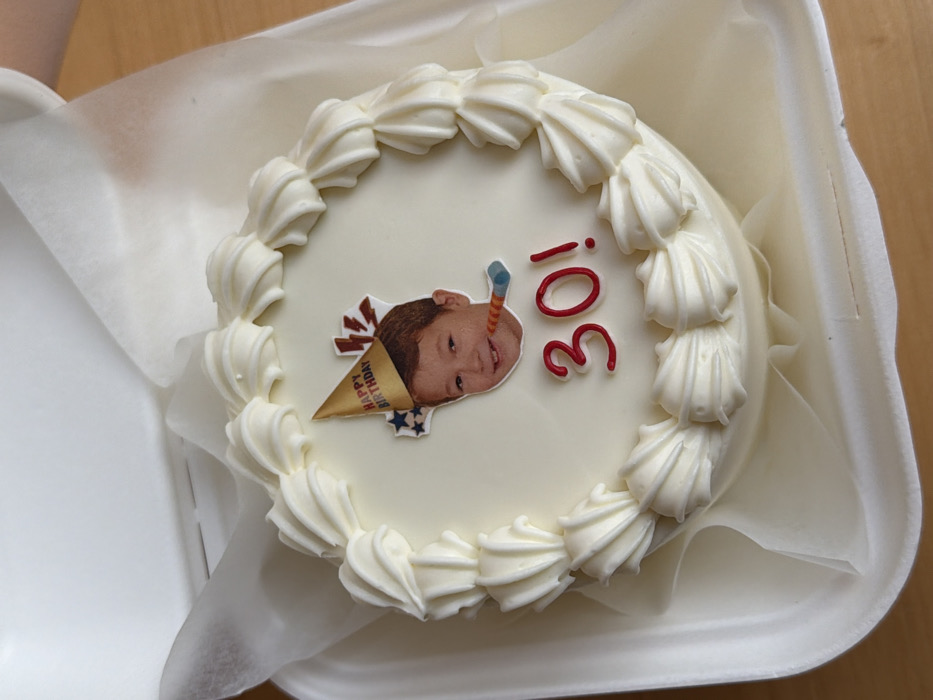 The scene depicts a round birthday cake, approximately 15-20 cm in diameter, resting inside an open white polystyrene container. The cake is covered in smooth, off-white icing, with a decorative border of piped cream-colored swirls encircling its top edge. Centered on the cake's surface is an edible image of a smiling child's face with light brown hair, appearing to wear a small gold "HAPPY BIRTHDAY" party hat and holding a striped party blower. Below the child's image, the numbers "30!" are written in red icing. The cake container is placed on a light-colored, possibly wooden, surface, showing a subtle grain. The illumination suggests an indoor setting. This scene is described as being in Tel-Aviv, Israel.