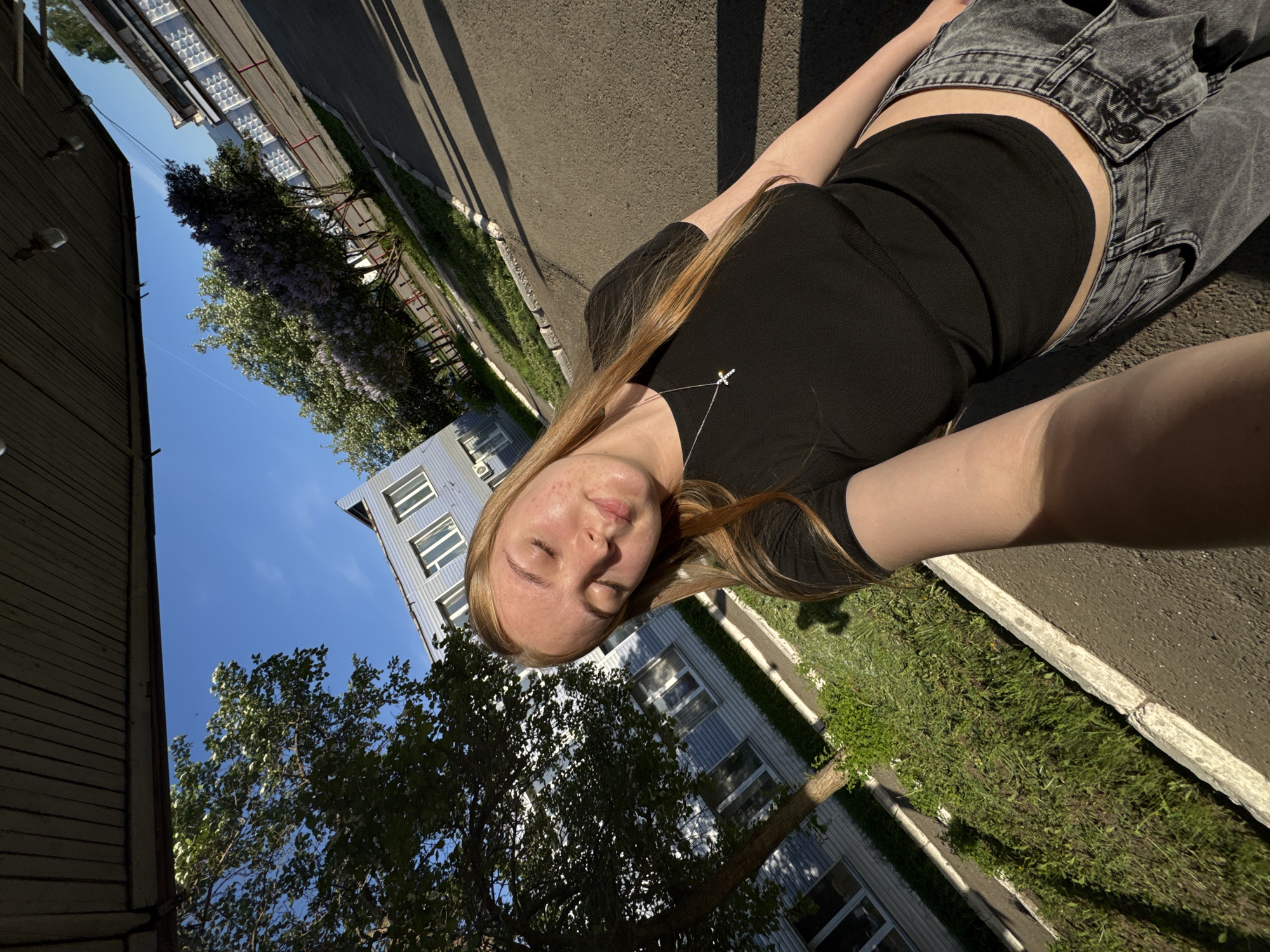 A young woman lies on a sidewalk next to a grassy strip. She wears a black top, grey jeans and a silver necklace with a small cross pendant. The photo is taken looking up at her, with buildings and trees in the background. The location appears to be Osa, Russia based on architectural details and general appearance.