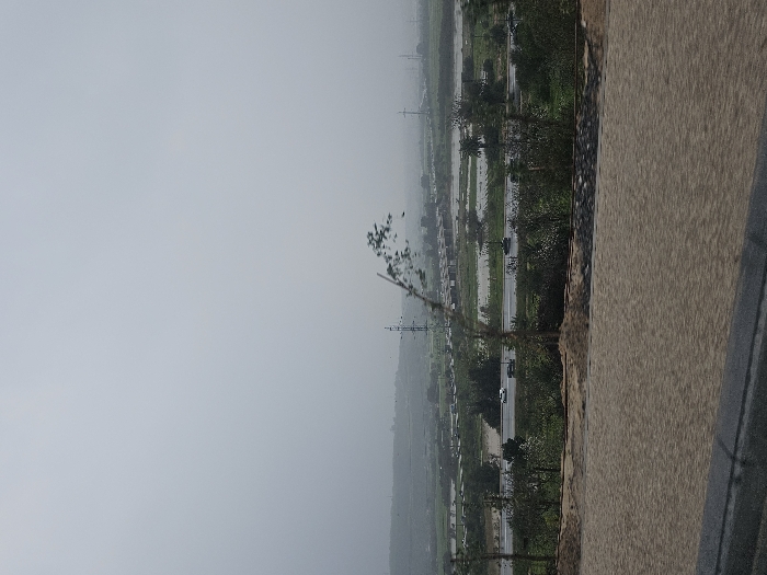 An elevated perspective reveals a landscape under a hazy sky. A multi-lane highway, intermittently dotted with moving vehicles, stretches through a verdant expanse. Patches of water reflect the sky amidst the greenery. In the foreground, a portion of a road or pathway is visible, suggesting the vantage point is from a higher location, possibly a bridge or elevated platform. Power lines punctuate the skyline in the distance.