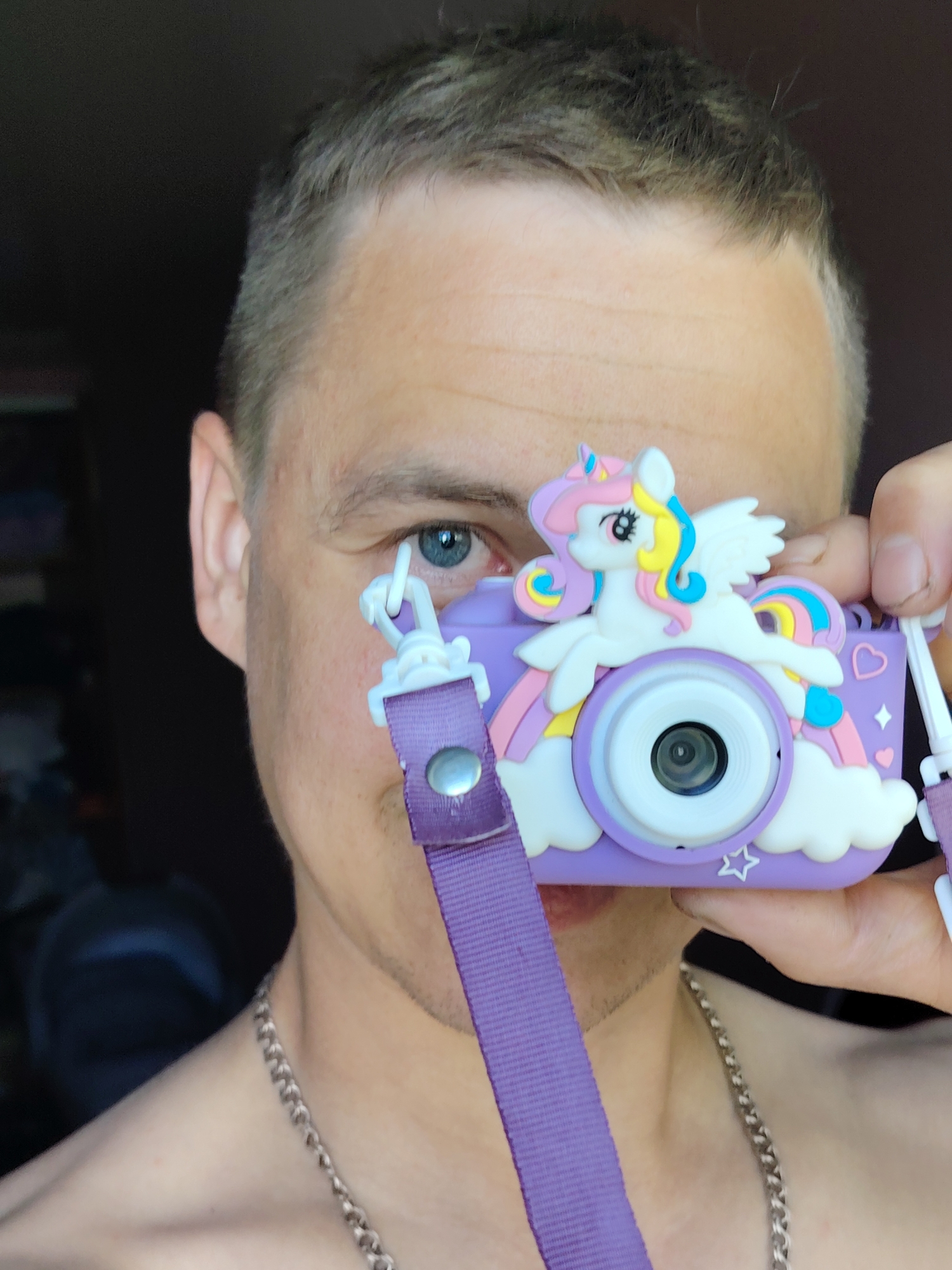 A medium close-up shot features a light-skinned individual holding a purple unicorn-themed camera up to their face. The camera obscures part of their face, but their left eye, forehead, and the left side of their face are visible. The individual has short, light brown hair and a silver chain necklace around their neck. The camera is adorned with a white unicorn figure, a rainbow, clouds, and stars. The camera strap is purple and has a silver snap. The background is dark and indistinct. It is not possible to determine the location of the image, including whether it is in the city of Chaikovsky, Russia.