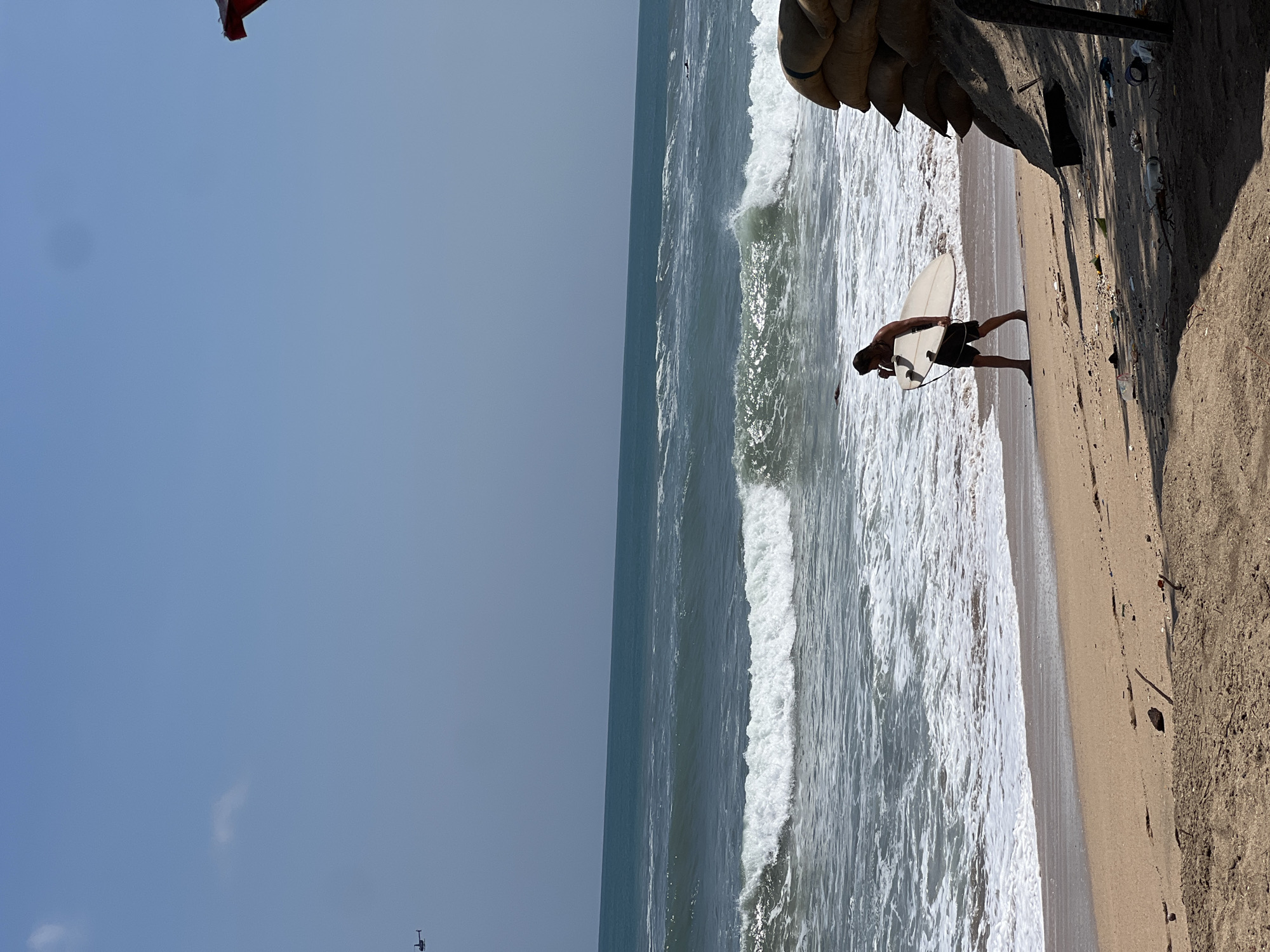 A surfer walks along the beach carrying a surfboard. The ocean has moderate waves, and the sky is clear. Sandbags are piled on the right side of the frame.