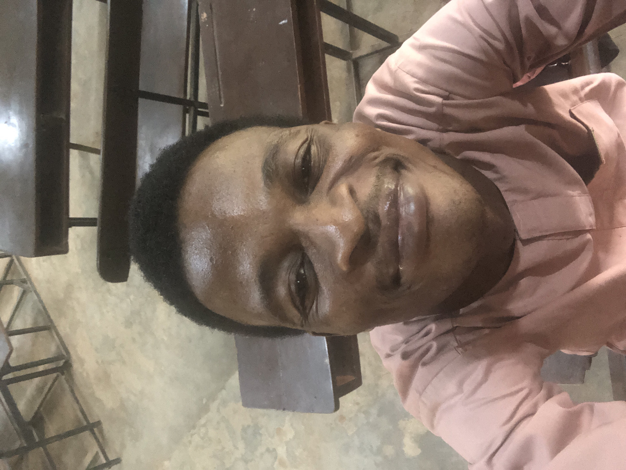 A man with dark skin and short, dark hair is taking a selfie in an indoor setting. He is wearing a pink garment with a collar and buttons. The man is smiling widely, and his face is illuminated by overhead lighting, creating highlights on his forehead and cheeks. The background consists of what appears to be wooden desks or benches with metal frames, suggesting a classroom or lecture hall. The floor is a light-colored, textured surface. The image is taken from a slightly upward angle.