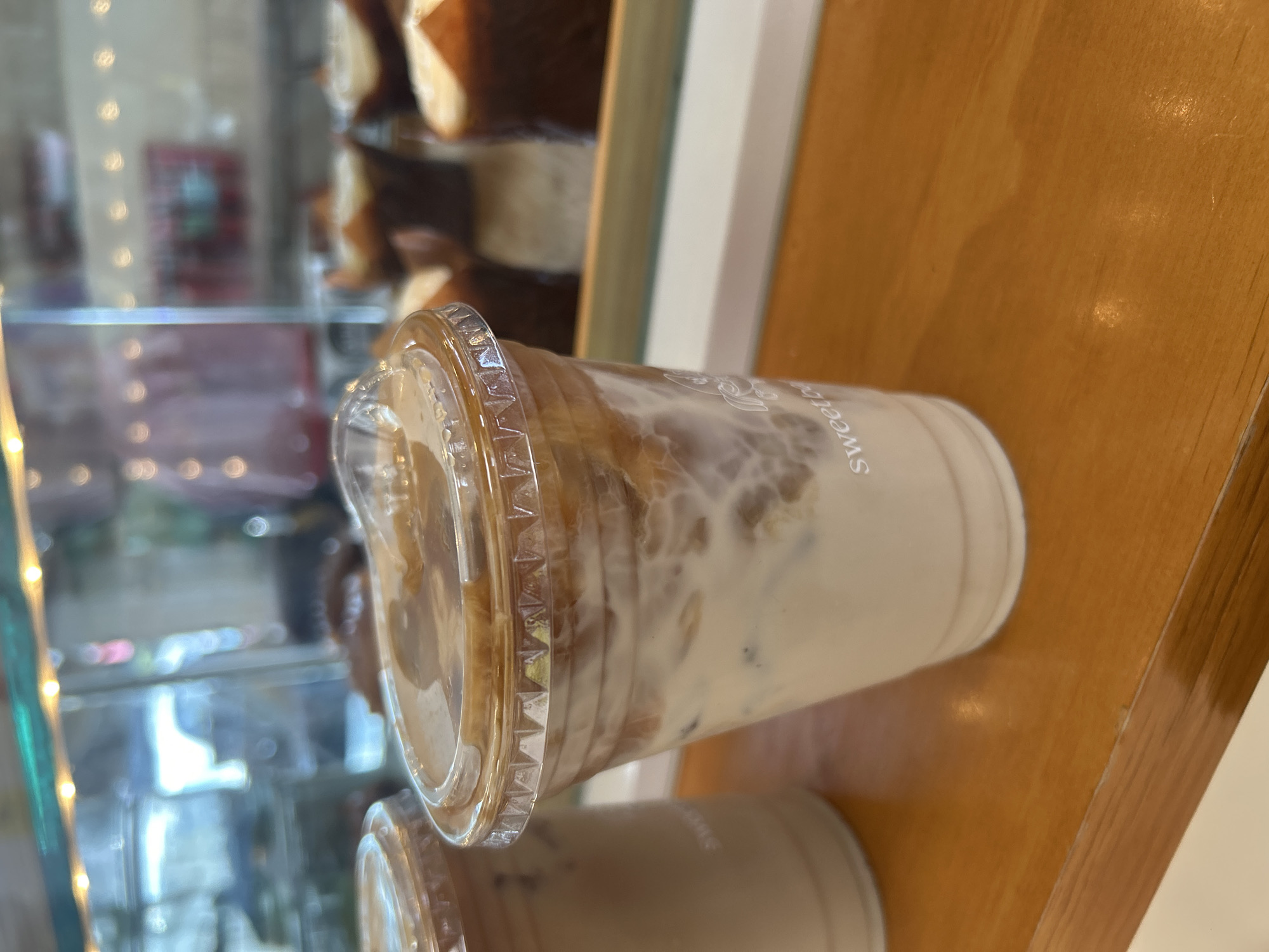 Here is a description of the image:
The image shows a plastic cup of iced coffee on a wooden surface. The cup has a clear lid and the drink appears to be a latte or similar coffee beverage with milk. Another cup of coffee is located behind it. In the background, there is a glass display case filled with baked goods, possibly sandwiches and pastries. It is difficult to establish the exact location or city.