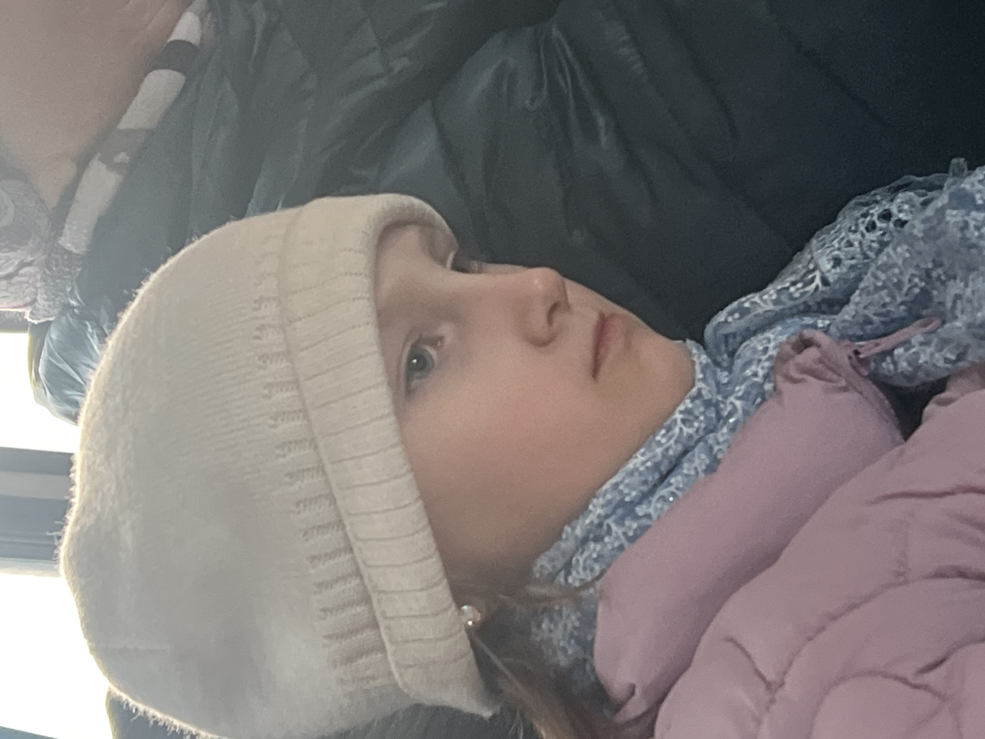 A young girl with fair skin, light eyes, and a light-colored knitted hat is looking towards the right side of the image. She is wearing a pink jacket and a blue patterned scarf. The background shows part of a dark-colored puffy jacket or coat. The location in the city of Noviy, Russia, cannot be determined from the image.