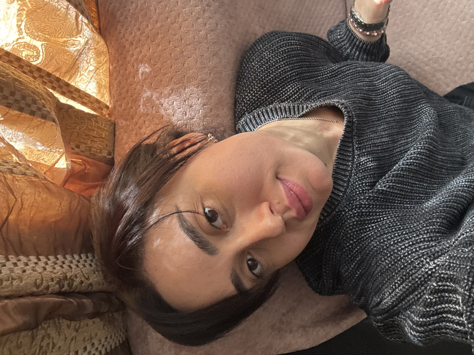 An adult female is depicted in a domestic interior within the city of Bolnisi, Georgia. The subject is positioned against a light peach-colored upholstered surface, wearing a dark grey ribbed knit sweater. She has dark brown, chin-length hair, brown eyes, and dark eyebrows, with a slight smile on her lips. Visible accessories include small, silver-colored hoop earrings on both ears, a thin silver-colored chain necklace, and multiple bracelets on her left wrist, one appearing dark and another beaded. In the background, behind the subject's head, draped curtains of a golden-brown or bronze fabric with an ornate pattern are visible, illuminated by a strong, warm light source, consistent with natural daylight from a window. The subject is looking directly into the camera, indicative of a self-portrait.