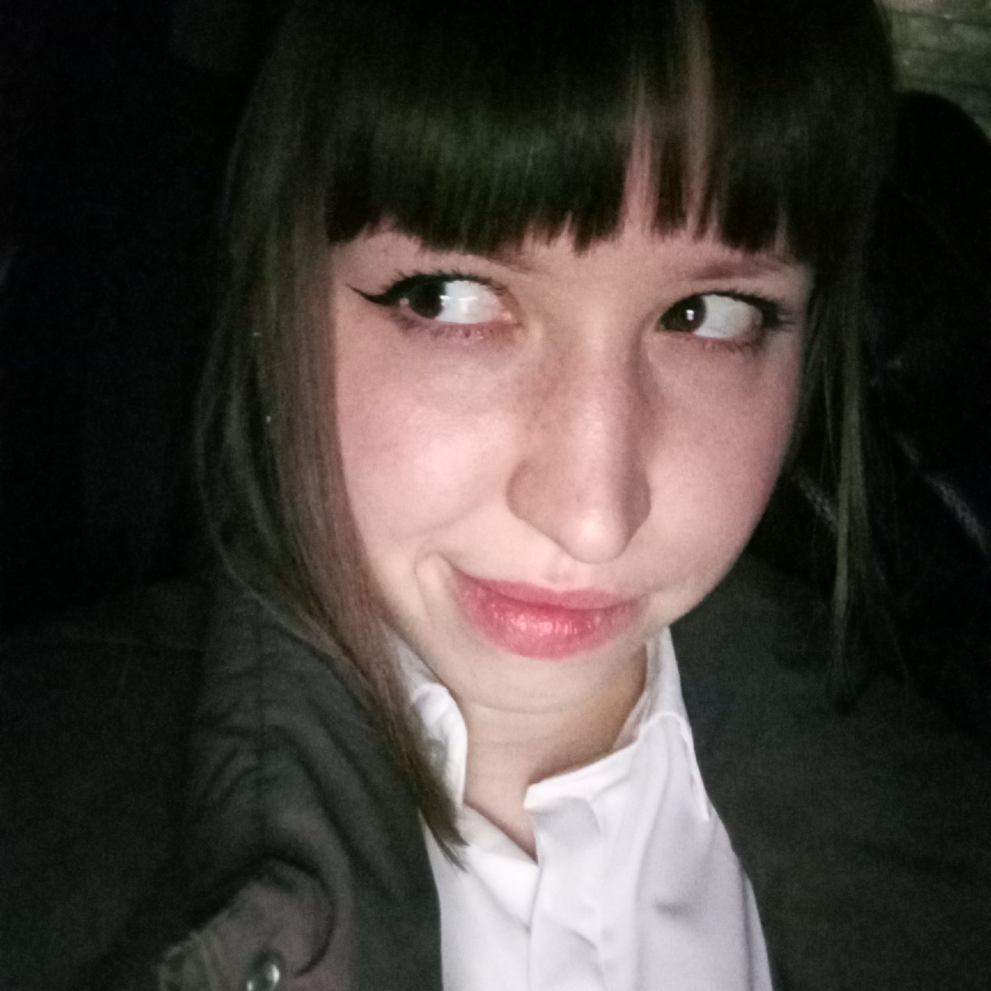 A woman with dark hair and bangs is looking to the side with a slight smirk. She has fair skin with freckles, and her eyes are lined with eyeliner. She is wearing a white collared shirt and a dark blazer. The background is dark and indistinct. The location is not discernible from the image.