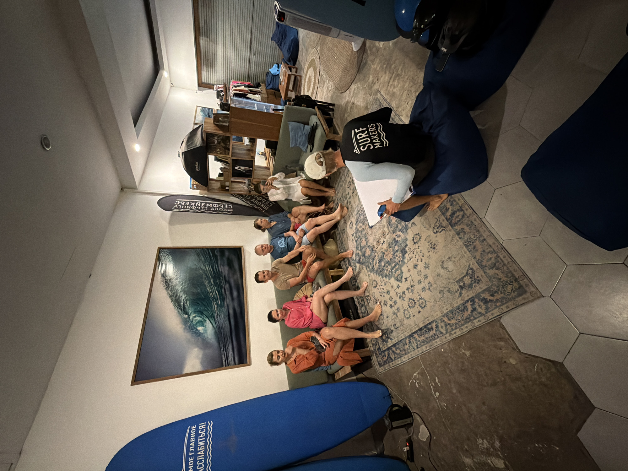A group of people sit on a couch in an indoor setting. A man in a "Surf Makers" shirt stands in front of them holding papers. The environment includes a surf board, a large picture of a wave, bean bag chairs, a rug, and shelves of items. This photo was likely taken in Kuta, Indonesia.