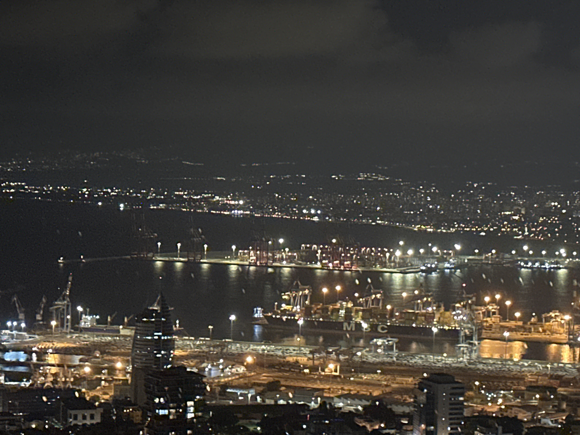 This is a nighttime aerial photograph of the port of Haifa, Israel. The image shows a large harbor filled with ships and cranes. The city lights are visible in the background, creating a sprawling urban landscape. The sky is dark and cloudy, with some lights visible from distant areas. The port is bustling with activity, as evidenced by the illuminated cranes and the lights on the ships. The overall scene depicts a major industrial and commercial hub in operation after dark.