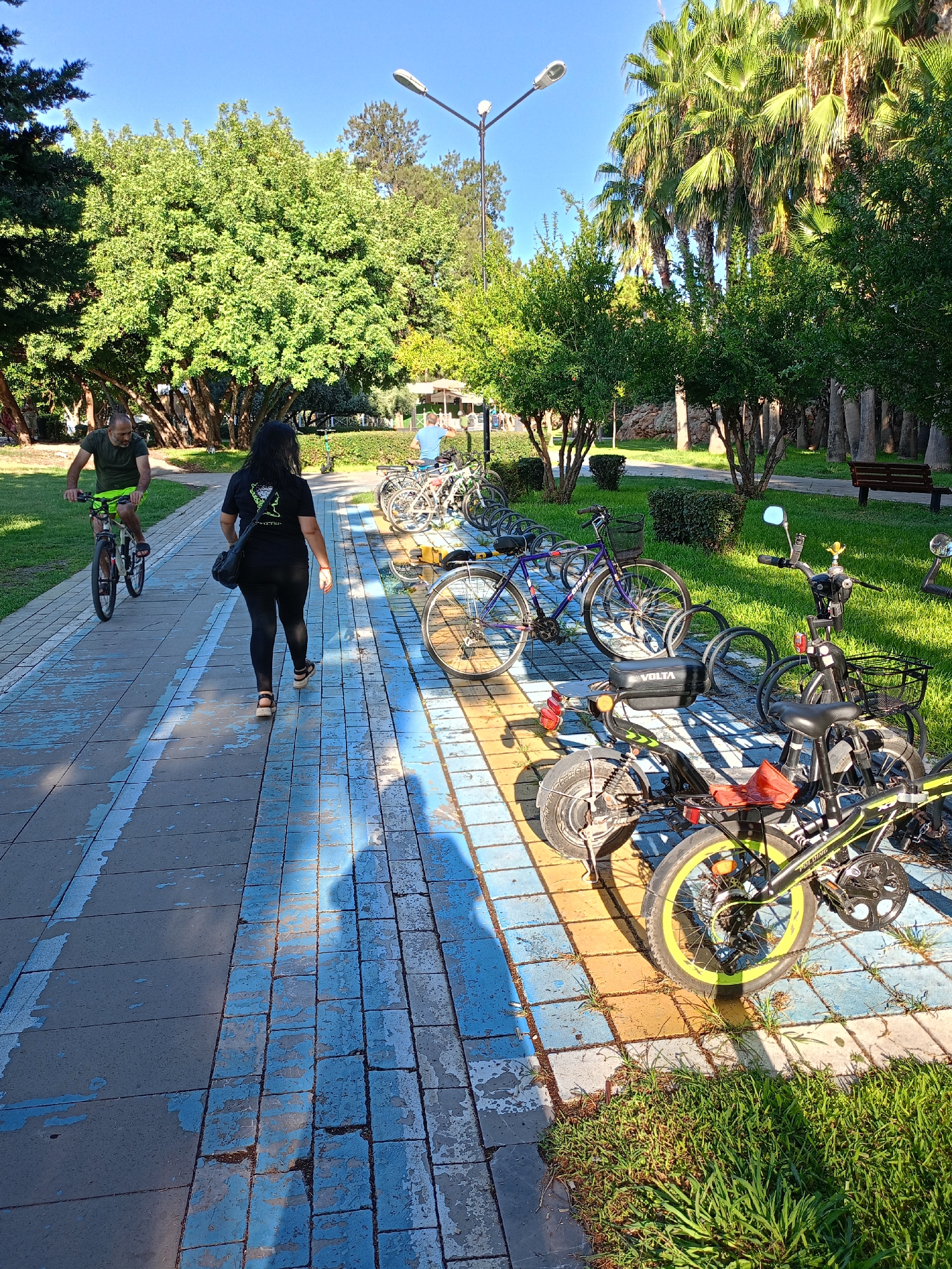 In Antalya, Turkey, a sunny day in a park setting shows a paved walkway with a blue and yellow painted pattern. A man wearing a dark green t-shirt and neon green shorts cycles on the left side of the path, casting a shadow. A woman in a black t-shirt and black leggings walks away from the camera on the same path, with a black bag slung over her shoulder. Several bicycles are parked in a rack on the right side of the path, with some leaning against the rack. One electric bike, black and green, is prominent in the foreground. Palm trees and lush green trees line the background, with a park bench visible in the far right. A streetlamp stands tall on the left side of the walkway. The ground is a mix of paved tiles and grassy areas.