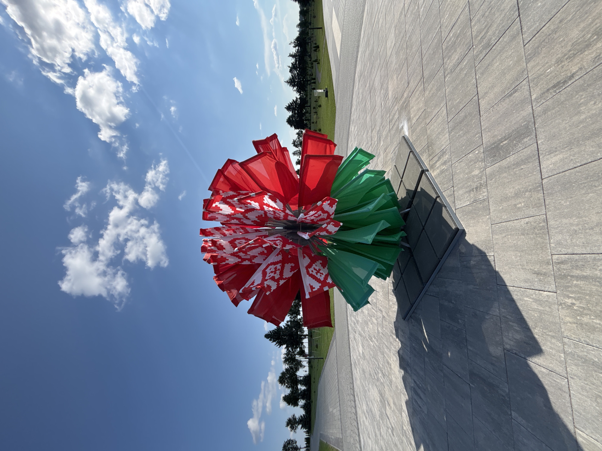 The image shows a decorative flower-like sculpture made of red and white flags with a Belarusian ornament and green flags at the bottom, displayed outdoors on a paved area. The sculpture is positioned in the foreground, with a clear blue sky and scattered white clouds above. In the background, there is a landscaped park area with trees, grass, paved pathways, and some lighting fixtures. The overall scene suggests a public space, possibly a park or memorial site, in the city of Ozyaritska-Slabadski rural council, Belarus. There are no people visible in the image.