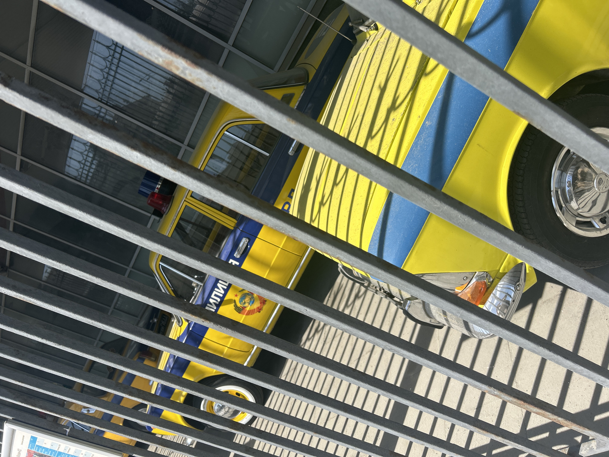 The image shows a yellow and blue Soviet-era police car seen through a metal railing. The car features the word "ПОЛИЦИЯ" (Police) written in Cyrillic and the Soviet Union coat of arms. The car has a red and blue emergency light on its roof. The surrounding environment appears to be an outdoor exhibition or museum, as indicated by the metal railing. The image is taken from a high angle, looking down at the vehicle. The location is Unknown, Unknown.