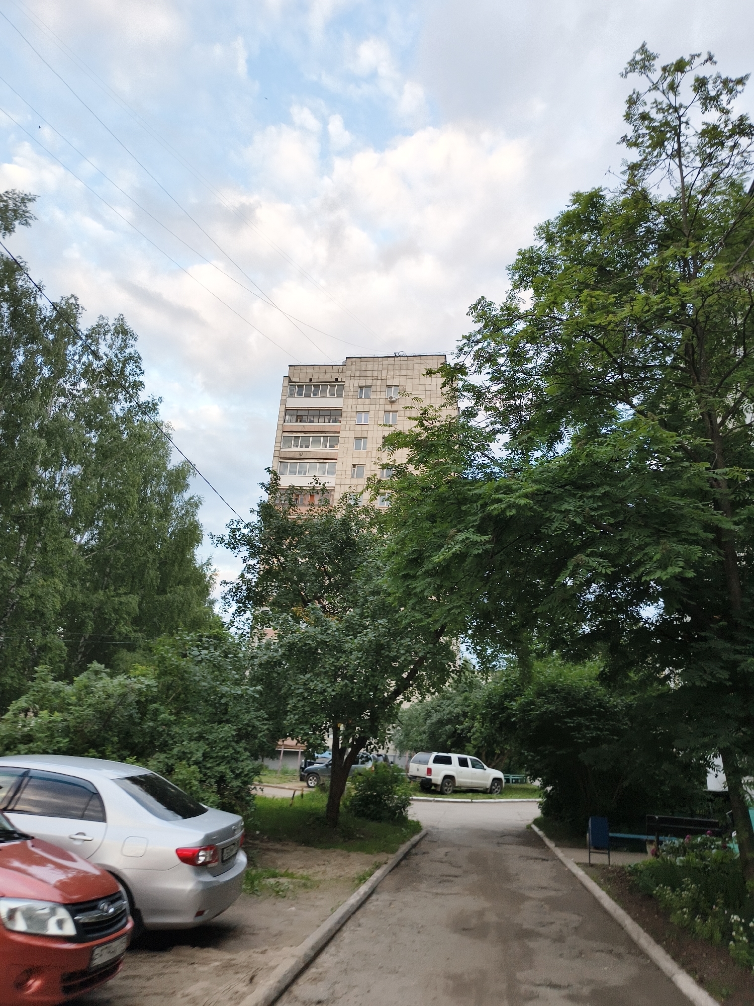The image shows a residential area with a multi-story building, trees, and parked cars. A tall, light-colored apartment building with numerous windows and balconies is visible in the background. Lush green trees with dense foliage surround the building and line the area. Several cars are parked on a dirt and asphalt path in the foreground. One car, a silver sedan, is parked on the left. In the middle distance, a white SUV is parked. The sky is cloudy with patches of blue. The scene appears to be a typical urban setting in Chaikovsky, Russia, likely a residential courtyard.