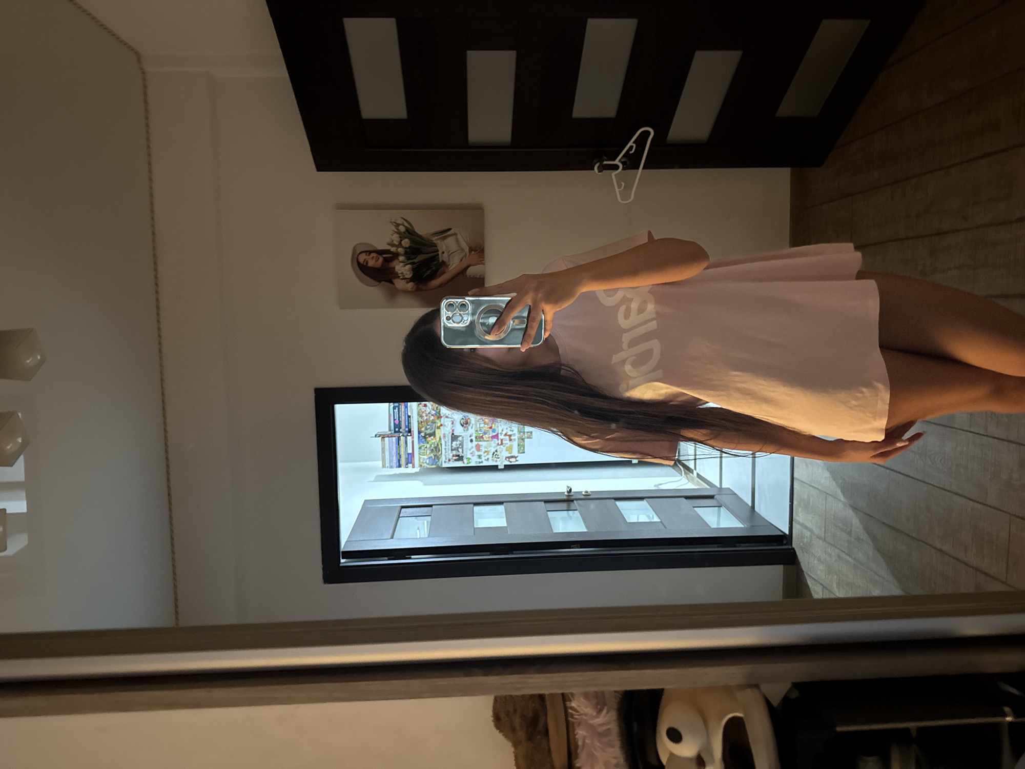 A young woman is taking a selfie in a bedroom, reflected in a large mirror. She is wearing a pink t-shirt dress with white lettering on the front. Her long, dark hair covers her face. In the reflection, a door with glass panels is visible behind her, along with a picture on the wall of a woman holding tulips. To the left of the mirror, a stuffed toy with large eyes and a black hat is partially visible. The floor is made of wooden planks. This appears to be an indoor residential setting.