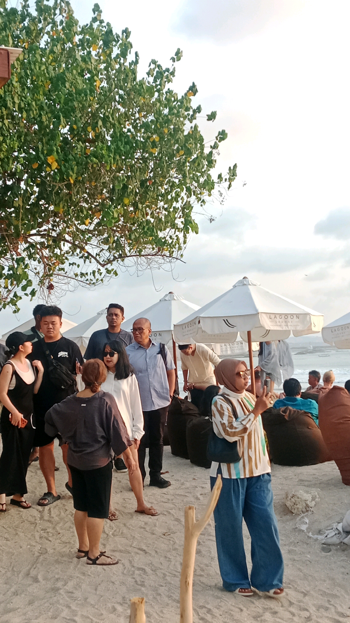This image depicts a beach scene in Kuta, Indonesia, likely during late afternoon or early evening. A group of people are gathered on the sandy shore, with a large tree providing shade in the upper left quadrant. In the background, white umbrellas, some branded with "LAGOON LOUNGE," are set up on the beach, suggesting a lounge or restaurant area. The ocean is visible in the distance.
Several individuals are the primary focus. In the foreground, a woman wearing a hijab and a striped shirt with wide-leg jeans is holding up a smartphone, appearing to take a photo or video. To her left, a woman in a dark top and black shorts is looking away from the camera. Beside her, a man in a black t-shirt and black shorts is also looking to his left. Behind them, a man in a light blue collared shirt and dark pants stands with his hands in his pockets. Another man, wearing a dark t-shirt, is positioned between the man in the blue shirt and the group on the left. Further back, other individuals are seen interacting or relaxing on beanbag chairs, some under the umbrellas. The sand is light-colored and appears somewhat damp in places. The sky is overcast with patches of bright light, indicating either approaching or receding daylight.