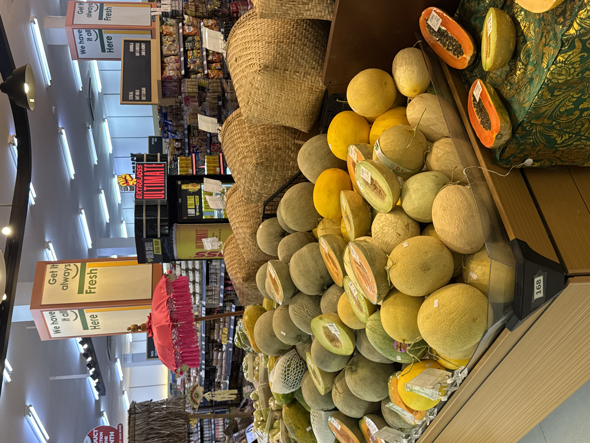 The image shows a display of fresh fruits, predominantly melons and papayas, in a grocery store. The fruits are arranged in a tiered display, with some cut open to reveal their interiors. Several large, woven baskets are placed behind the fruit display. In the background, shelves stocked with various grocery items are visible. Overhead, fluorescent lights illuminate the store. Two signs are prominently displayed: one reads "We have it all Here" and the other "Get it always Fresh". Another sign in the background advertises "EXTRAJO55 ULTIMATE". The setting appears to be a supermarket in North Kuta, Indonesia, suggested by the display of local fruits and the general aesthetic of a Southeast Asian market.