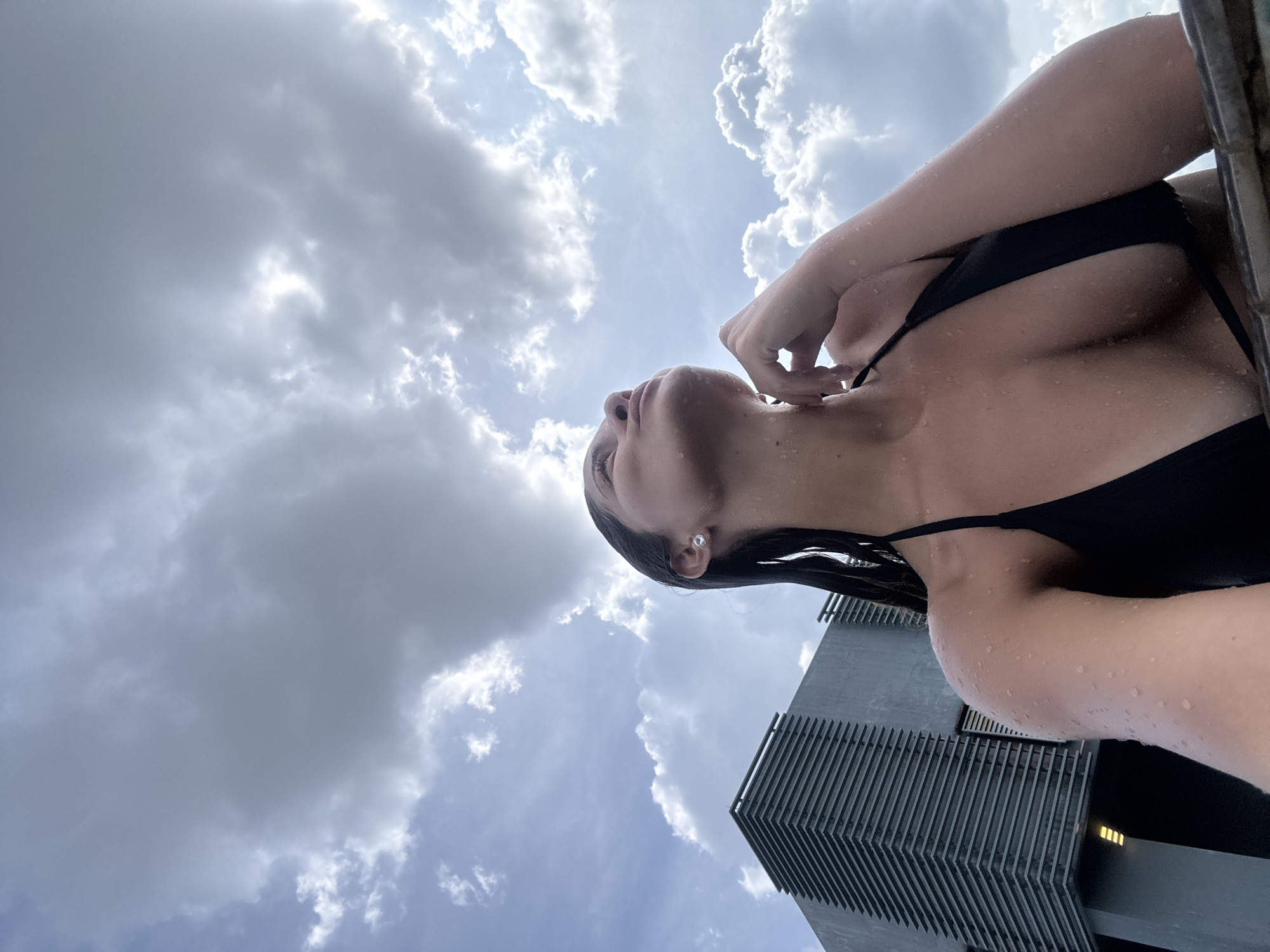This low-angle photograph captures a person looking upwards against a dramatic sky, set in the urban environment of Ampang Jaya Municipal Council, Malaysia. The main subject is a female, seen from the chest up, wearing a black bikini or swimsuit top. Her skin and wet dark hair are adorned with numerous water droplets, indicating she has recently been in or near water, possibly a pool. Her head is tilted back and her gaze is directed towards the sky, with one hand gently touching her neck or collarbone area.
The sky occupies the majority of the frame, showcasing a mix of bright white, sunlit clouds and darker grey clouds, suggesting a partly cloudy but bright daytime. Patches of lighter blue sky are visible between the cloud formations. On the right side of the image, a segment of a modern building with a distinctive dark, slatted or ribbed facade rises sharply, providing an architectural element to the scene. The perspective makes the building appear tall and imposing. The overall impression is one of serene contemplation or relaxation in an outdoor, possibly poolside or rooftop setting, under a vast and dynamic sky. There is no visible text in the image.