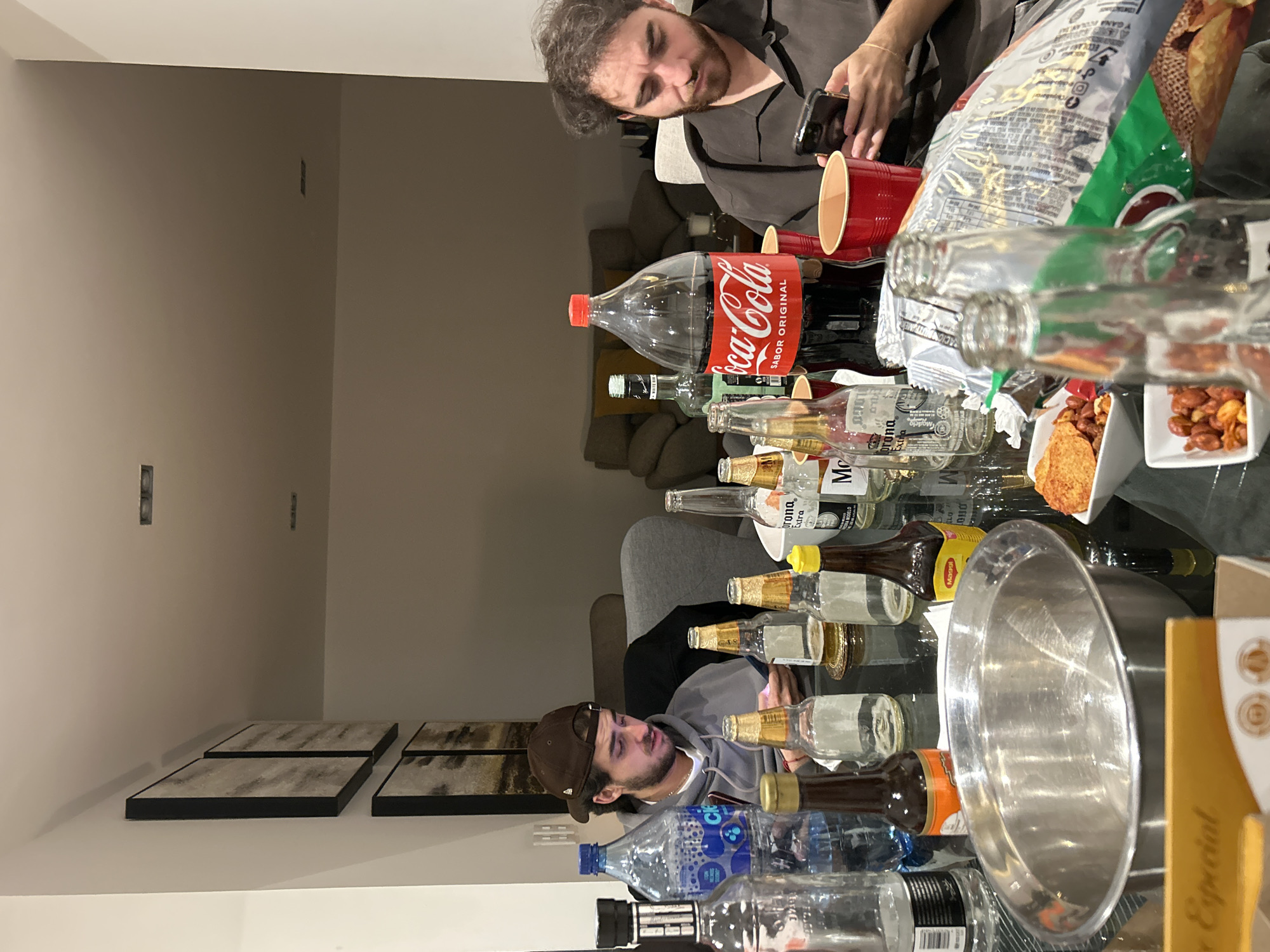 The image depicts two men sitting at a table cluttered with empty beer bottles (Corona, Modelo), a large Coca-Cola bottle, snack bags, and bowls of nuts. One man is looking at his phone, while the other is visible from the side, also looking at a phone. The scene appears to be indoors, likely in a home setting, with paintings on the wall and a couch visible in the background. Based on the OCR results, the scene is likely located in Mexico, possibly near a restaurant, given the "Especial" tag. Specific city cannot be verified with the image alone.