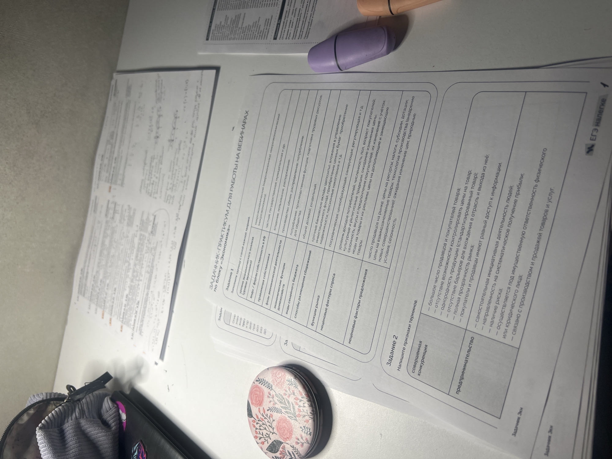 The image shows a workspace cluttered with papers, a vape pen, and other personal items, suggesting someone is working on a school assignment. The primary focus is on two sheets of paper with Russian text, titled "ЗАДАЧНИК-ПРАКТИКУМ ДЛЯ РАБОТЫ НА ВЕБИНАРАХ" which translates to "WORKSHOP-PRACTICUM FOR WORKING IN WEBINARS", related to the subject of "Economics". The sheets contain tables and prompts, indicating homework or study material. Other items include a grey purse/bag and a small, round container with a floral pattern. The lighting is bright and the background is a plain, off-white surface. The location is not discernible from the information in the picture.