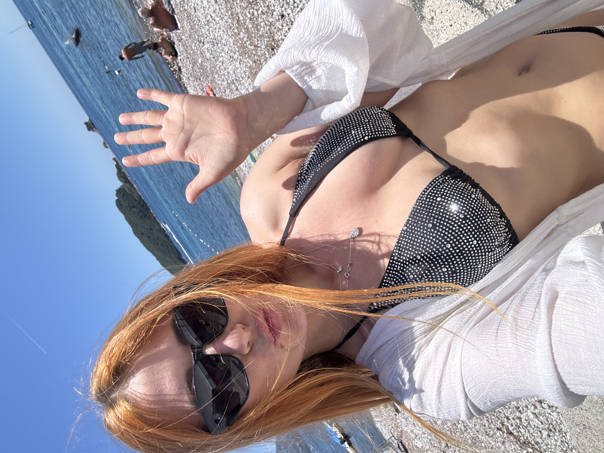 An overhead, slightly tilted shot captures a woman relaxing on a pebbly beach in Budva, Montenegro, under a clear blue sky. The main subject is a woman with long, reddish-blonde hair, wearing dark sunglasses, a black bikini top adorned with small shimmering details, and a loose, sheer white cover-up. She is lying down, with her right hand raised towards the camera, palm open. A delicate silver necklace with a pendant is visible around her neck, resting on her chest.
The bright sunlight indicates a clear, sunny day, likely mid-day or afternoon, casting gentle shadows across her body. In the background, the turquoise-blue sea stretches out, where several other people are visible swimming or wading. The shoreline is made of small, light-colored pebbles, and distant green hills or mountains line the horizon across the water. The scene portrays a relaxed, leisurely summer day by the sea. No text is visible in the image.
