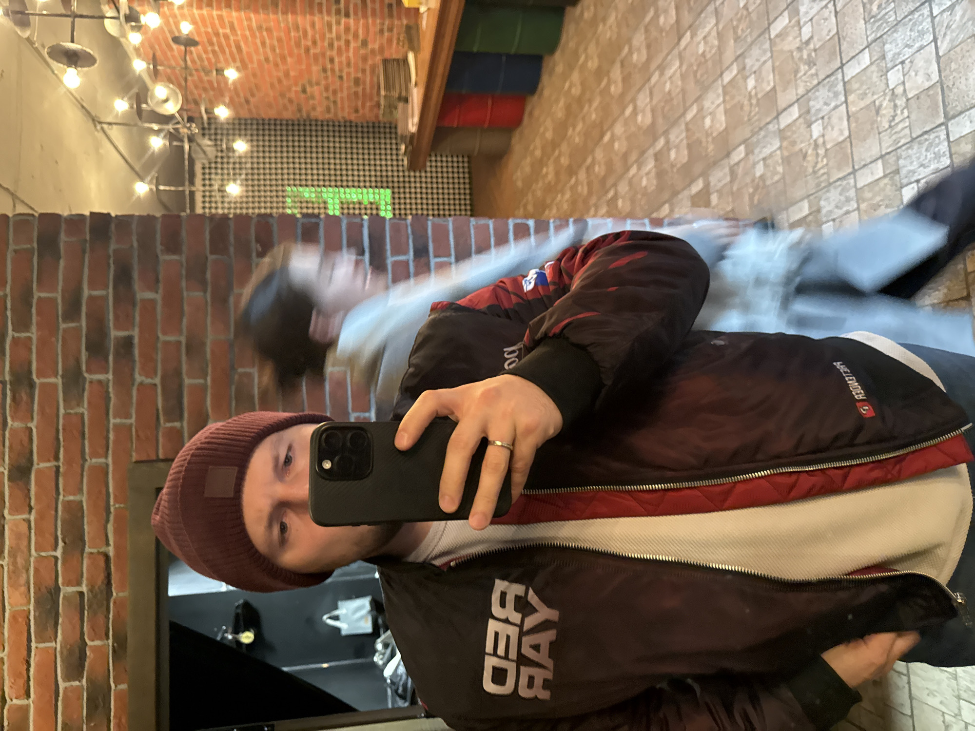 The image appears to be a selfie taken in a mirror within a commercial establishment.
Subject: The primary subject is a person wearing a maroon beanie, a white shirt, and a black and maroon bomber jacket with "RED RAY" visible in mirrored text. The person is holding a black smartphone, presumably to take the picture. A wedding ring is visible on their ring finger.
Environment: The background includes a brick wall, an assortment of hanging light fixtures, and decorative elements such as shelves with colorful containers. The floor is tiled. A blurred figure is reflected in the mirror behind the person taking the selfie.
Location (deduced from jacket details): Based on the "RED RAY" emblem on the jacket, it is highly likely that this photo was taken in Krasnoyarsk, Russia. "RED RAY" is a store that is located in this city.