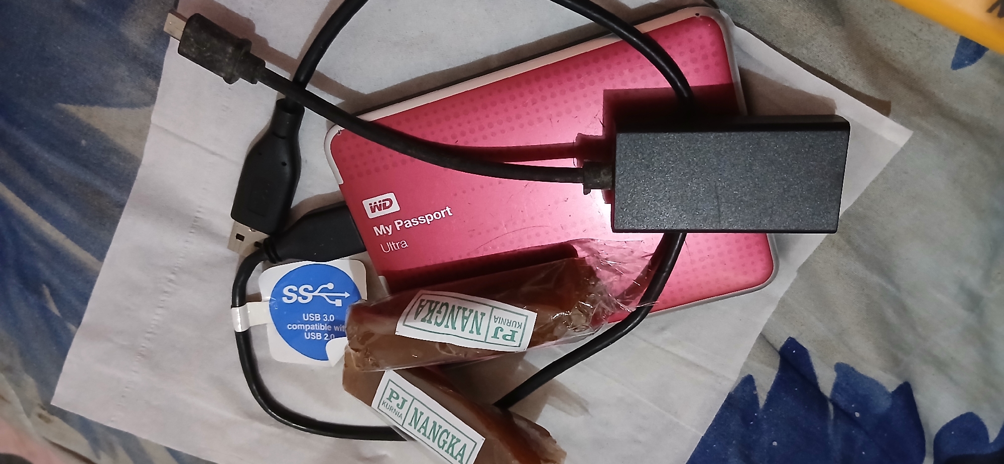 The image shows a close-up, high-angle view of a pink Western Digital My Passport Ultra external hard drive and its associated cables, along with two individually wrapped food items. The hard drive, which appears to be a 2TB model based on its size and common configurations, is connected to a USB cable. A small black adapter is also visible, attached to one of the cables. The food items are cylindrical and wrapped in clear plastic, with white labels bearing the text "PJ NANGKA" and "KURNIA". A blue sticker with "SS" and "USB 3.0 compatible with USB 2.0" is also attached to a cable. The objects are resting on a white tissue or paper napkin, which is itself placed on a surface with a blue and white patterned fabric. The lighting suggests an indoor setting, possibly a room in a residential or commercial building. The image does not contain any specific details to indicate a location within the city of Samarinda, Indonesia.