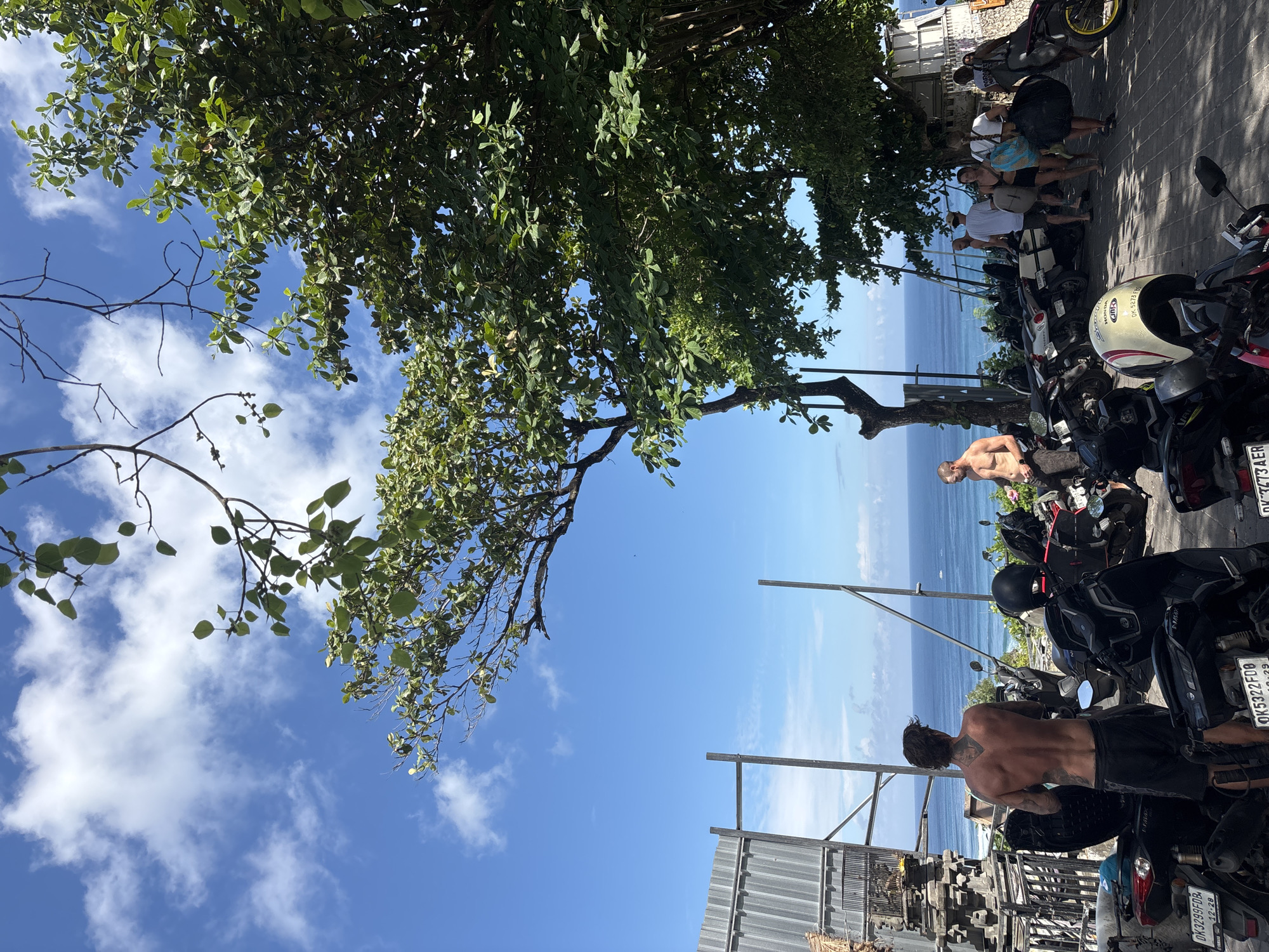 The image depicts an outdoor scene in Kuta Selatan, Indonesia, with a partial view of the ocean framed by trees, a sky with scattered clouds, and various objects. The primary focus is on a parking area filled with parked motorbikes. Several people are present, including shirtless men near the motorbikes. To the left, there are construction or temporary structures, and in the distance, there is a group of people standing near parked motorbikes. The scene seems to be taken during the day under bright sunlight.