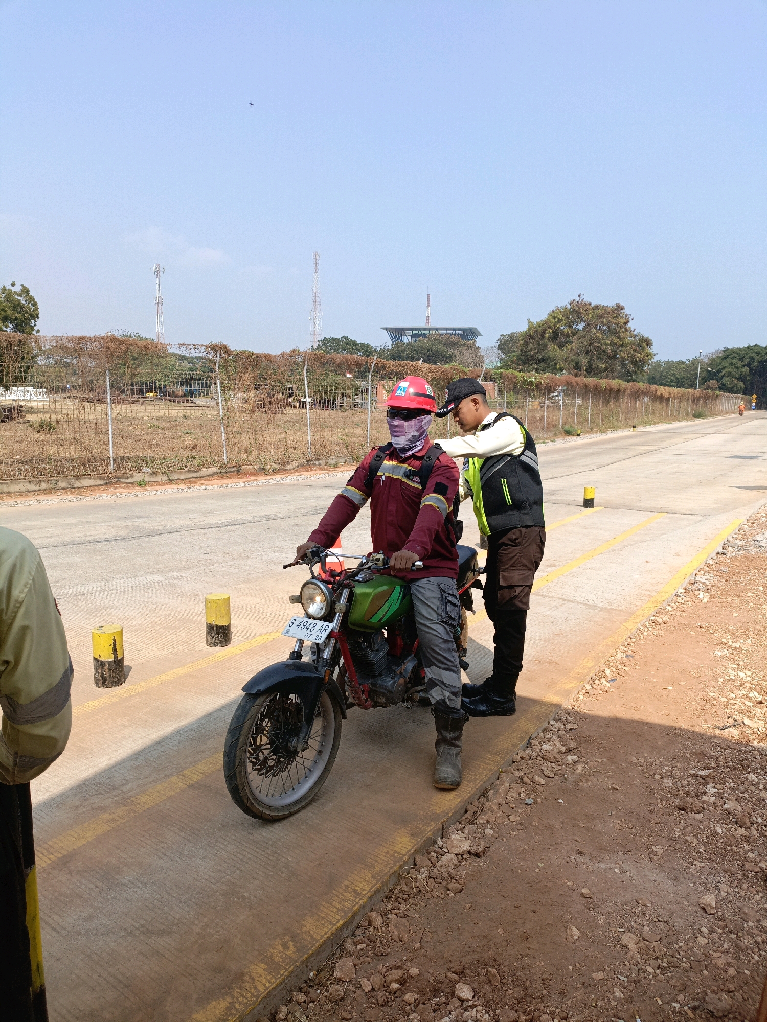 In Jenu, Indonesia, two men, possibly workers, are interacting near a motorcycle on a paved road. The man on the motorcycle wears a red helmet, a maroon long-sleeved shirt with yellow accents, gray pants, and gray boots. He has a bandana or cloth covering his lower face. The second man, standing beside the motorcycle, is wearing a black cap, a white and yellow safety vest over a black shirt, and dark pants with brown accents. He appears to be talking to or instructing the man on the motorcycle, his hand resting on the other man's shoulder. The motorcycle is green and black with a visible license plate reading "S 4948 AR" and "07 28". Yellow and black striped bollards are present on the road's edge, and a yellow line is painted on the pavement. The environment suggests an industrial or construction area, with a chain-link fence and dry vegetation in the background. Several transmission towers are visible in the distance against a clear blue sky. The ground on the right side of the road is unpaved and covered with dirt and rocks.