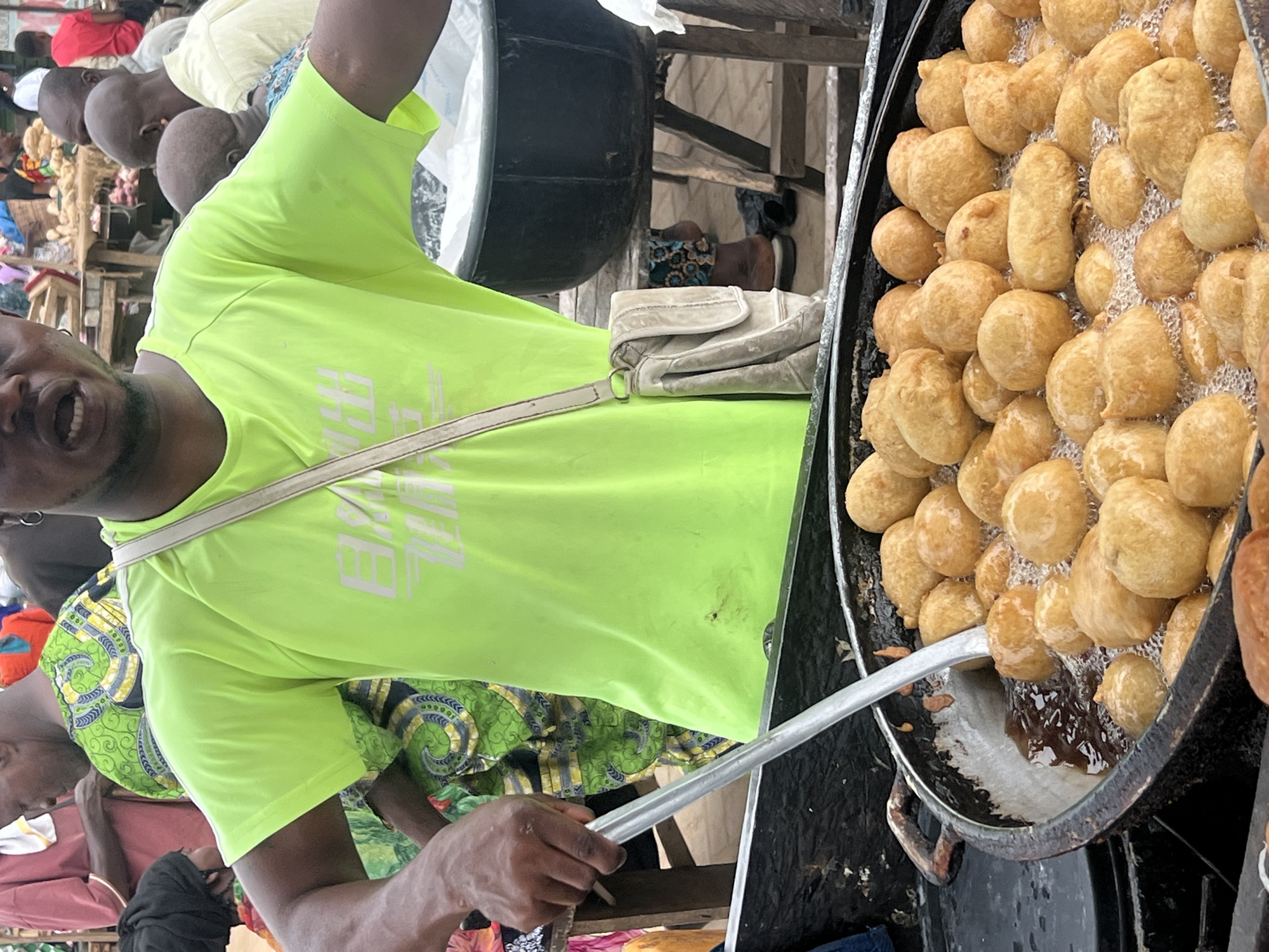 The image shows a man in Egan, Nigeria, preparing fried dough balls in a large, black wok filled with hot oil. The man is wearing a bright green t-shirt and holding a large, metal spoon. He appears to be flipping or stirring the dough balls in the oil. A small, white shoulder bag is visible near the man's waist. The surrounding environment suggests a busy, outdoor market or street food setting with other people nearby.