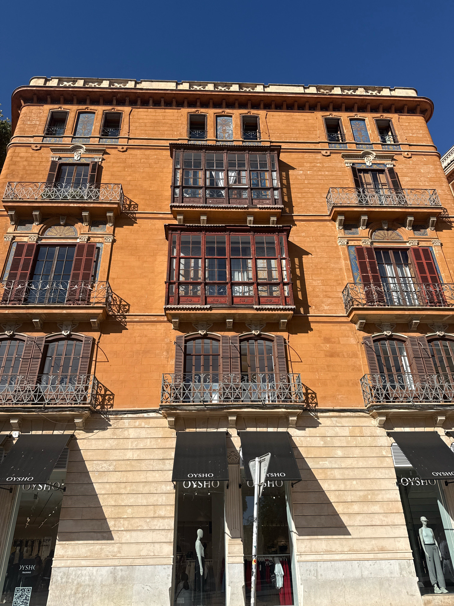 This is a low-angle, full-frame shot of an orange-colored, multi-story building on a sunny day, likely in Palma, Spain, given the architectural style and the "OYSHO" branding visible on the ground floor storefronts. The building features a classic architectural design with several floors of windows and balconies.
The upper floors exhibit ornate details, including decorative tiling around some windows and intricate wrought-iron railings on the balconies. The windows on these upper floors are mostly closed, some with wooden shutters partially drawn. Several projecting bay windows with dark wooden frames and glass panes are prominent features.
The ground floor of the building is occupied by OYSHO retail stores. Large glass windows reveal mannequins displaying clothing and racks of garments. Black awnings with the OYSHO logo are extended over the storefronts. The building's facade is made of brickwork on the upper levels, transitioning to a lighter stone material on the lower levels where the shops are located. The sky above is a clear, deep blue. A street sign and a traffic light are visible in front of one of the storefronts.