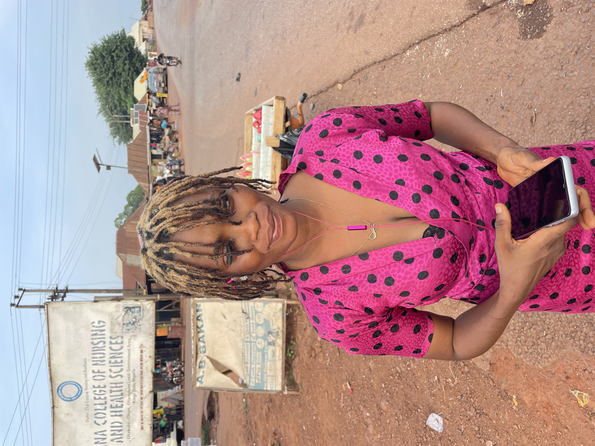 A woman stands on a dirt road in what appears to be Apunu, Nigeria, based on the sign visible in the background. She is wearing a pink dress with black polka dots and holding a smartphone. Her hair is styled in blonde dreadlocks. She also wears headphones and a necklace with a pendant. The road and surrounding area appear dusty and unpaved.
Behind her is a sign that reads "IINA COLLEGE OF NURSING AND HEALTH SCIENCES" along with an address and contact information. Another sign reads "A. B BAKAR" with a list of financial services. In the background, there are buildings, people, and motorcycles, suggesting a busy street or marketplace.