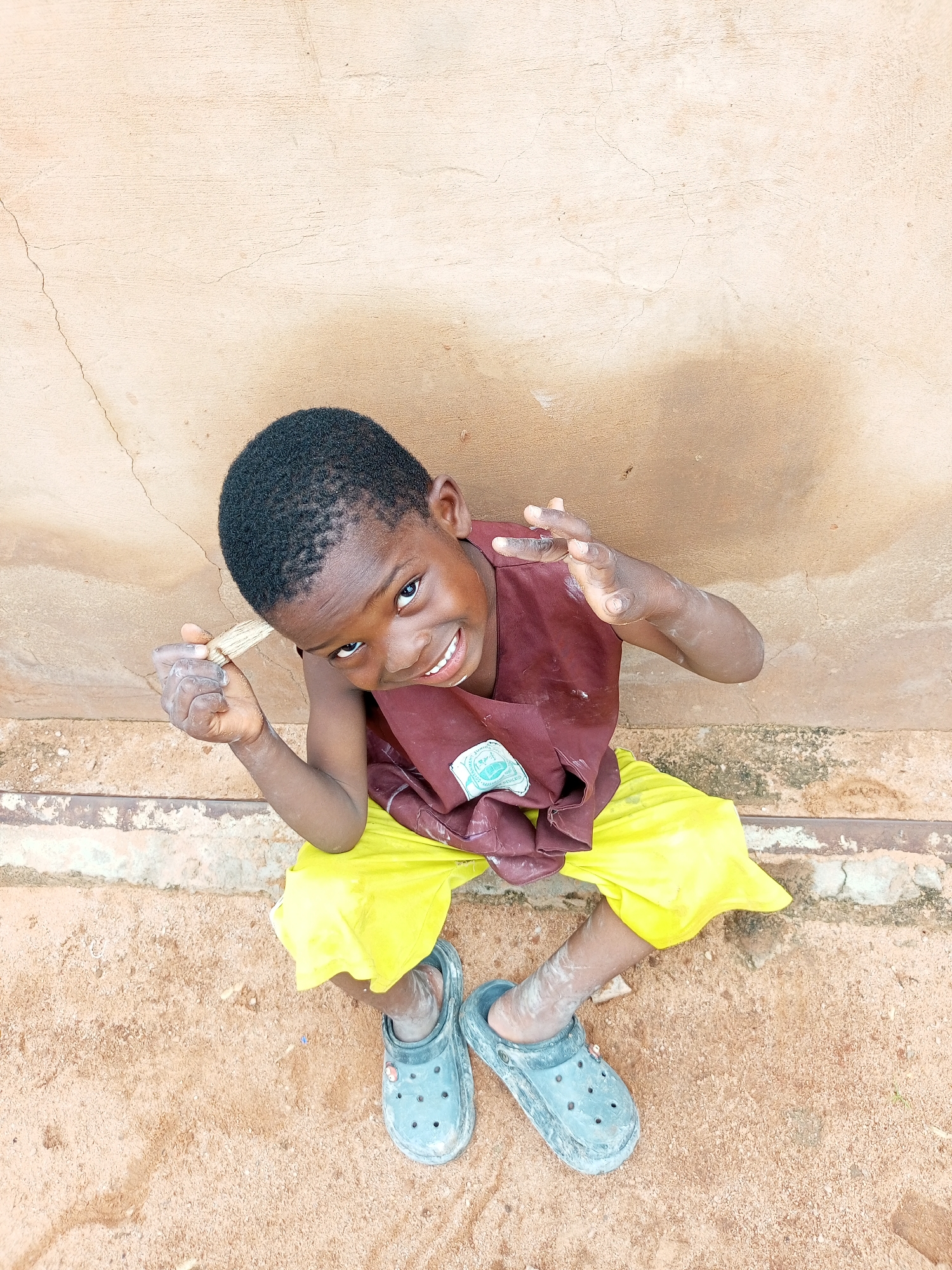 A high-angle, full-shot captures a young Black boy sitting on a dirt ground in front of a light brown, stucco wall in Tempure, Nigeria. The boy is wearing a maroon t-shirt, bright yellow shorts, and grey Croc-style shoes. He is smiling and looking up at the camera, with his right hand raised and holding a piece of wood. His left hand is also raised. White chalk dust is visible on his hands and arms, as well as on his right shoe. The wall behind him has a prominent crack running vertically. The ground is composed of reddish-brown dirt and small stones. The scene appears to be outdoors.