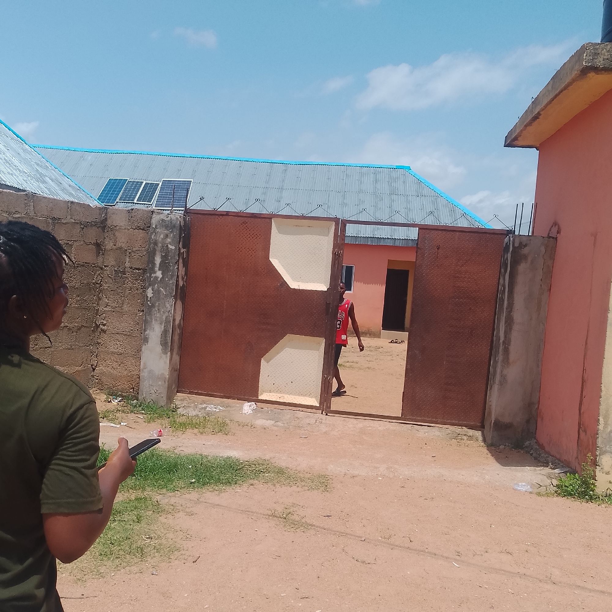 In Jalingo, Nigeria, on a clear, sunny day, a young person with dreadlocks, wearing a dark green t-shirt, stands facing away from the camera and looking at their phone. Behind them, a large, reddish-brown metal gate is partially open, revealing a pathway and a building. A young man wearing a red basketball jersey with the number '3' and black shorts is walking through the gate. The scene is set in an outdoor area with a dusty, sandy ground, patches of green grass, and a brick wall on the left. A corrugated metal roof with blue trim is visible on a building in the background, with solar panels mounted on it. To the right, a portion of a salmon-colored building with a concrete ledge is visible. The overall impression is a casual, everyday scene in a residential or commercial area.