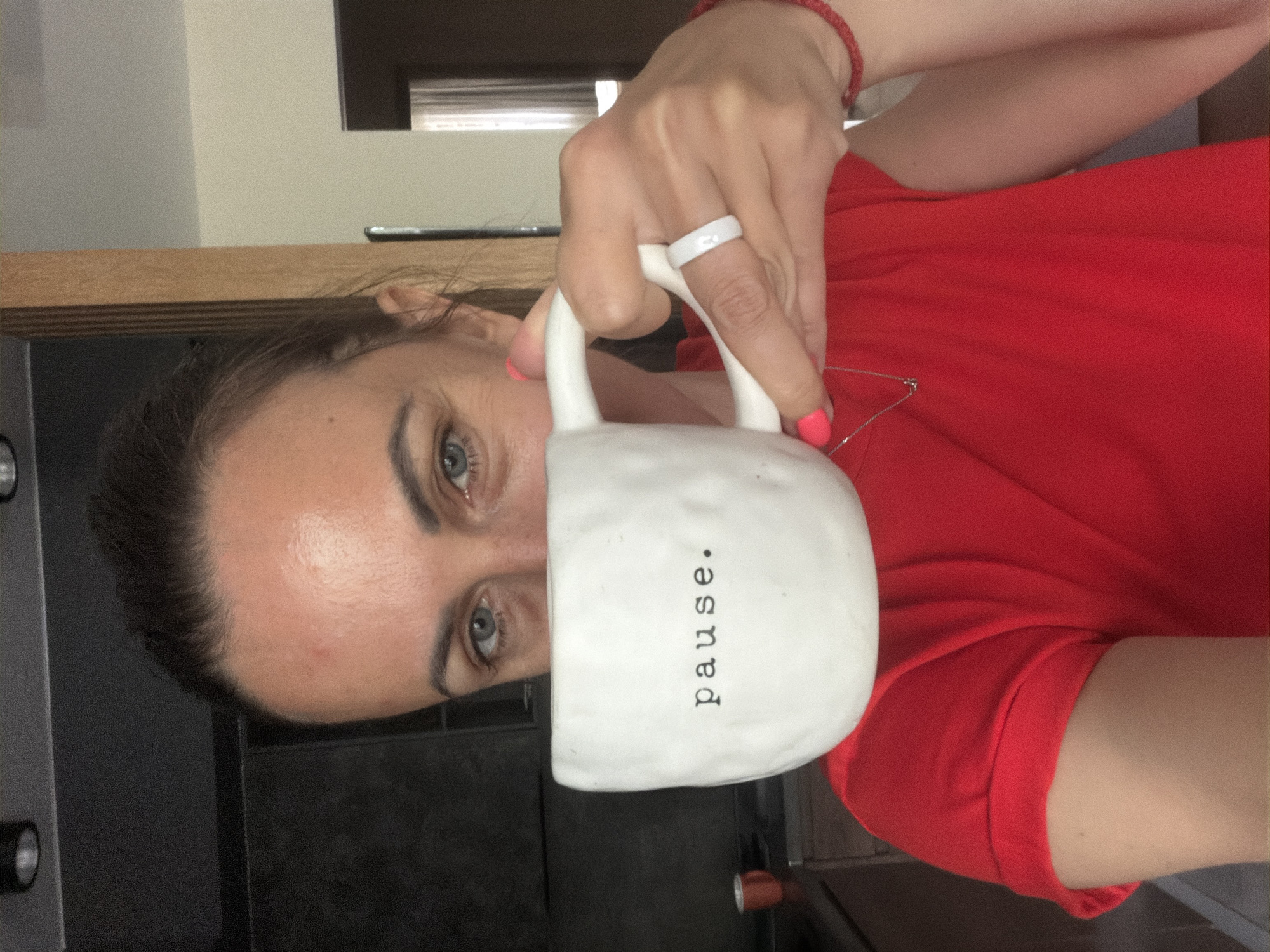 A woman is holding a white mug with the word "pause." printed on it. She is wearing a red shirt, a white ring on her finger, and a red bracelet on her wrist. Her fingernails are painted pink. She is standing indoors. The location is not discernable, and there is no evidence to suggest that the location is Birobidzhan, Russia.