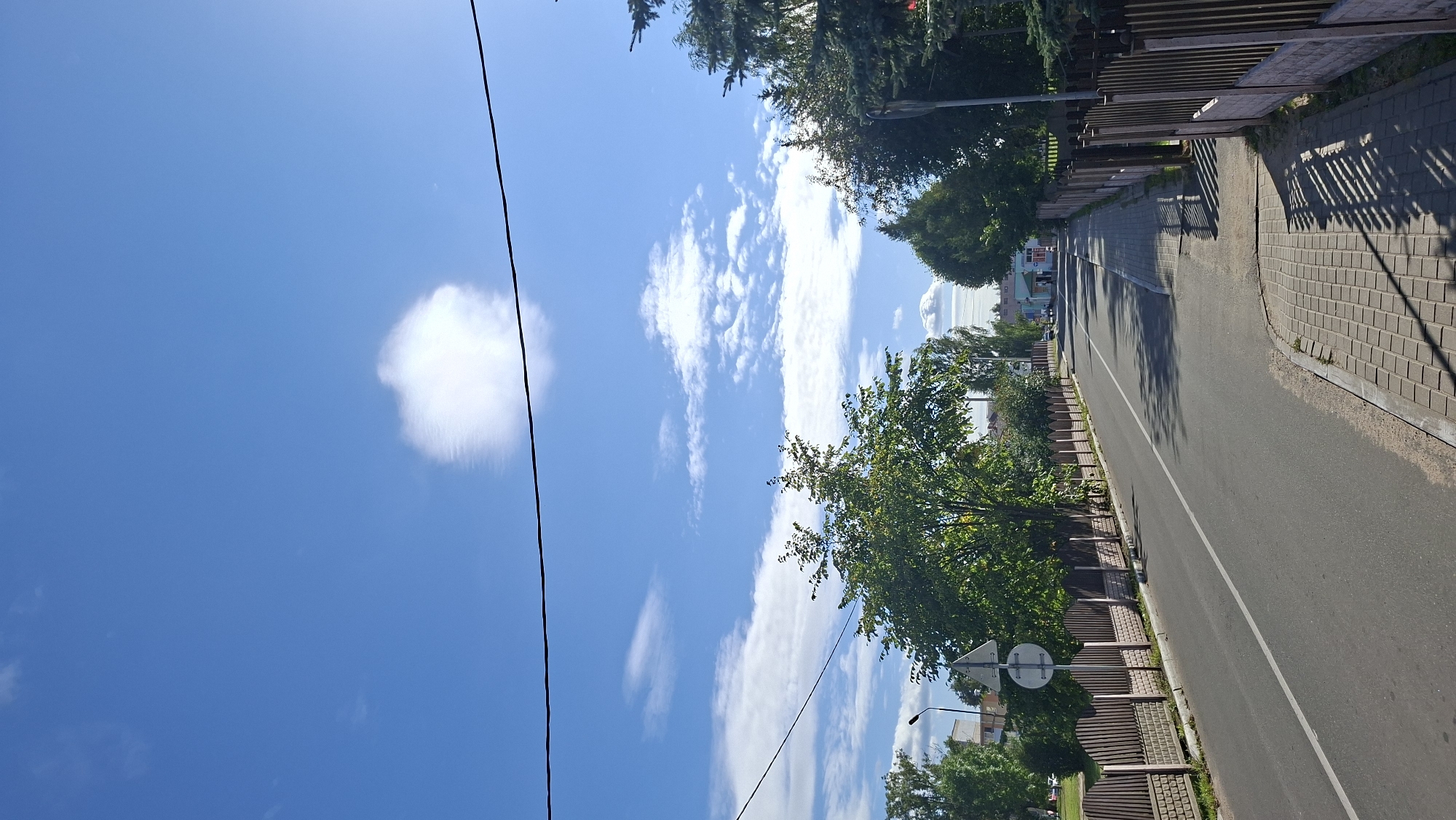 This is a vertical, eye-level, full-shot photograph of a street in Myadzyel, Belarus. The sky is a bright, clear blue with a few wispy white clouds. A single black power line stretches horizontally across the top of the frame. The street is asphalt with a white line down the center. On the left, a paved sidewalk runs alongside a wooden fence. Beyond the fence, there are trees and some buildings. On the right side of the street, there is a brick and concrete sidewalk, also bordered by a wooden fence, with trees beyond it. The sun casts long shadows across the sidewalk from the fence. The street appears to be in a residential or semi-rural area.