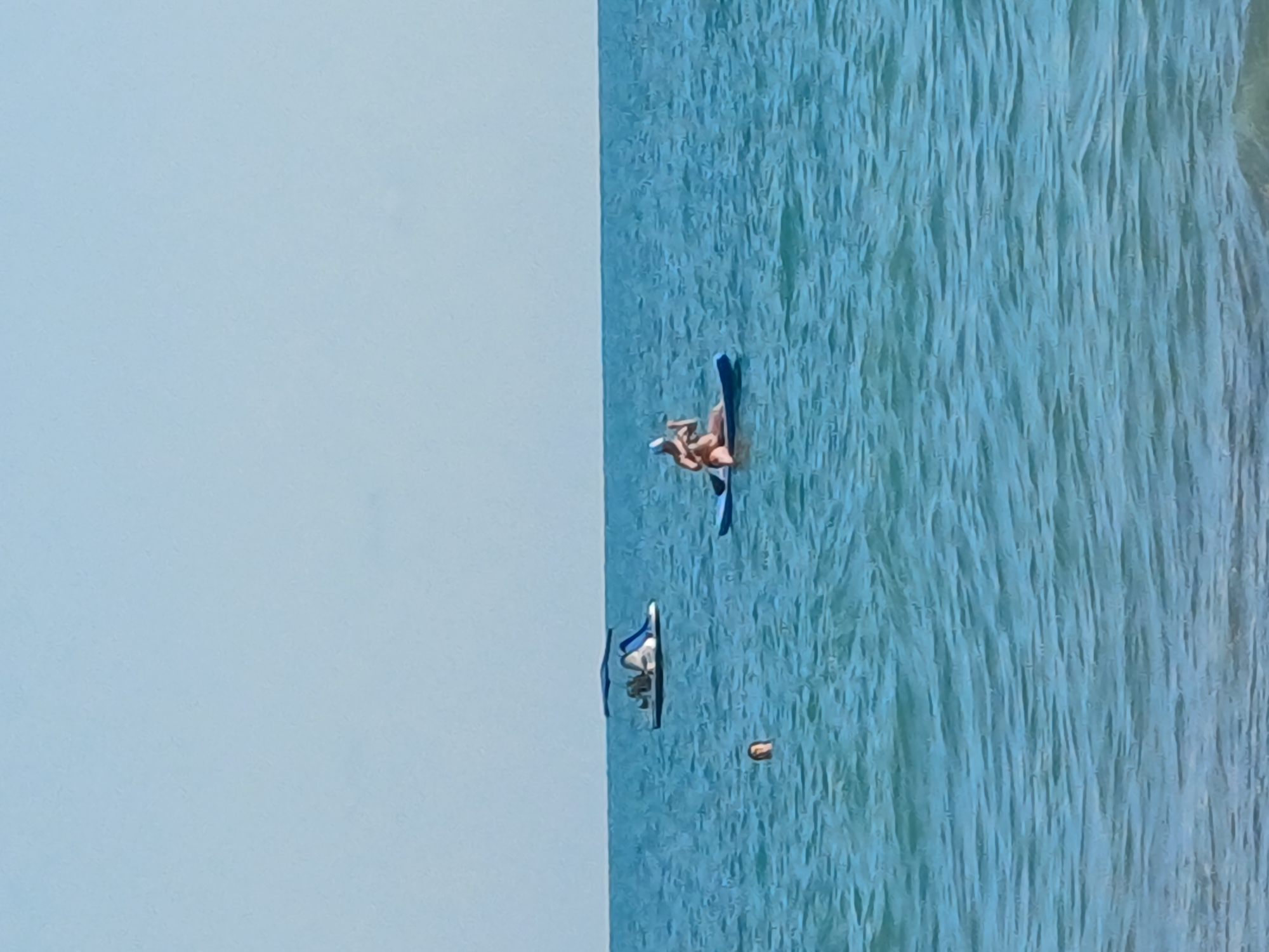 The image depicts a bright, sunny day at a beach in Sochi, Russia. The scene is dominated by the turquoise blue waters of the sea, with gentle waves creating a textured surface. In the water, two individuals are engaged in stand-up paddleboarding. One person is lying on their paddleboard, appearing relaxed and sunbathing, while another is visible on a separate paddleboard further down. The sky is a clear, pale blue, suggesting fair weather. The overall atmosphere is one of leisure and enjoyment of water sports on a pleasant day.