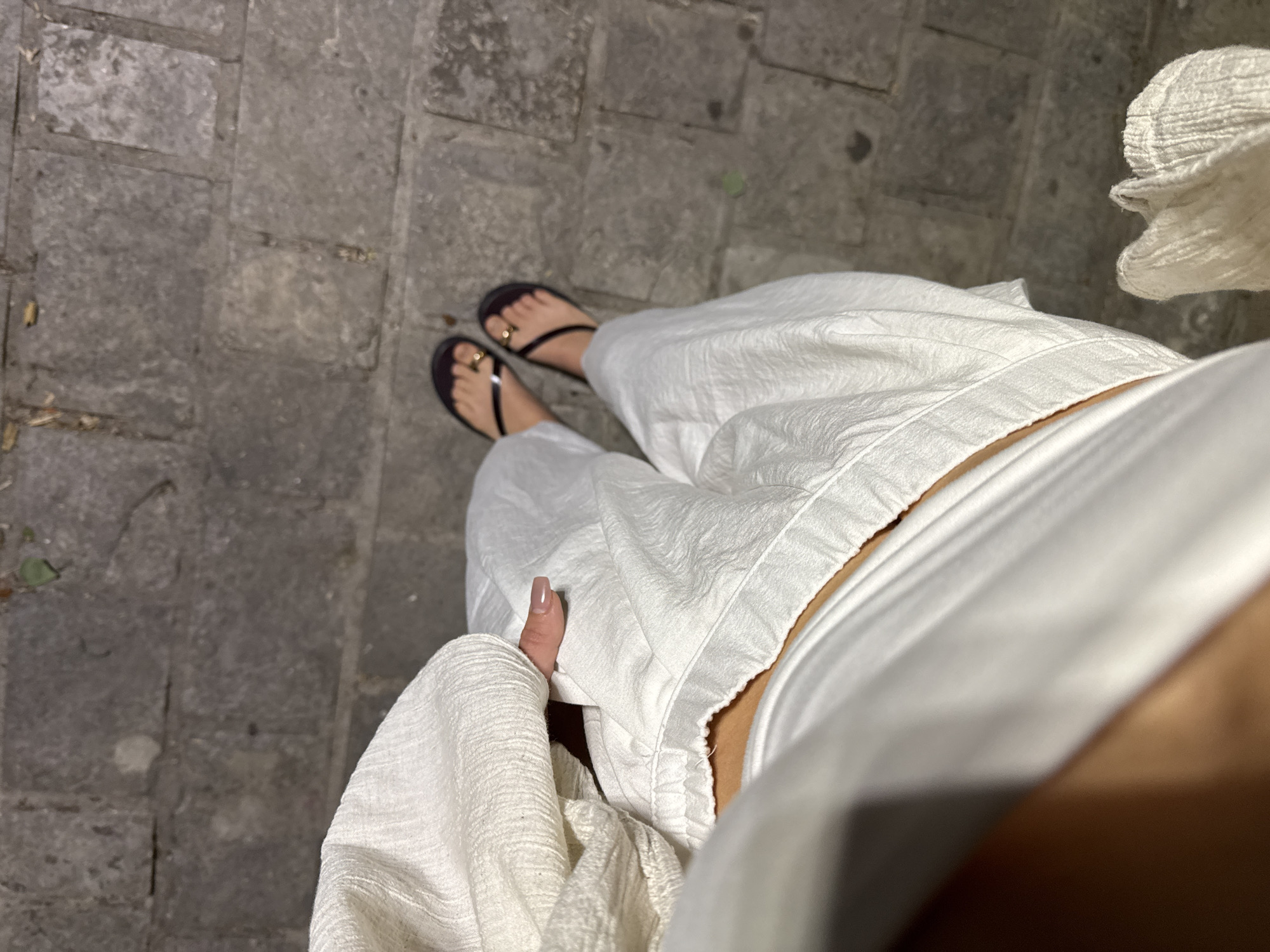 A person's legs are shown from a high-angle perspective, standing on a cobblestone path. They are wearing white wide-leg pants and dark flip-flops with gold toe rings. The left hand, with manicured nails, rests on the white pants. The cobblestone path appears to be in an outdoor urban setting, possibly in Budva, Montenegro, given the typical architecture of the area.