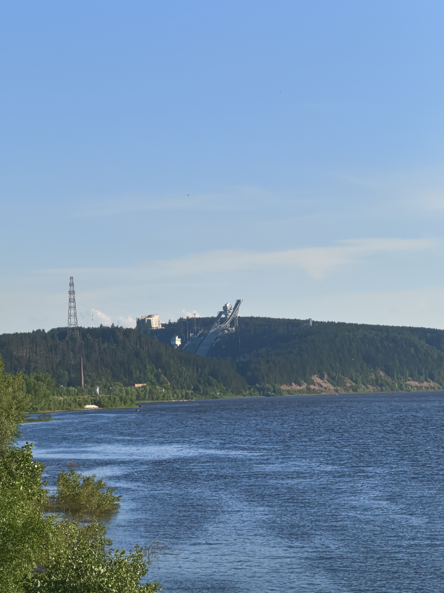 An aerial view shows a wide, blue river with ripples on its surface. On the right side of the river, a steep, forested hill rises, with a sandy cliff face near the water. At the top of the hill, a large, complex industrial structure with a ramp-like feature is visible, along with several buildings and a tall lattice tower. The sky is clear and blue with a few wispy clouds. The left foreground of the image features lush green trees and foliage along the riverbank. This scene is likely located in Olkhovka, Russia, possibly near a ski jumping or industrial complex overlooking the river.