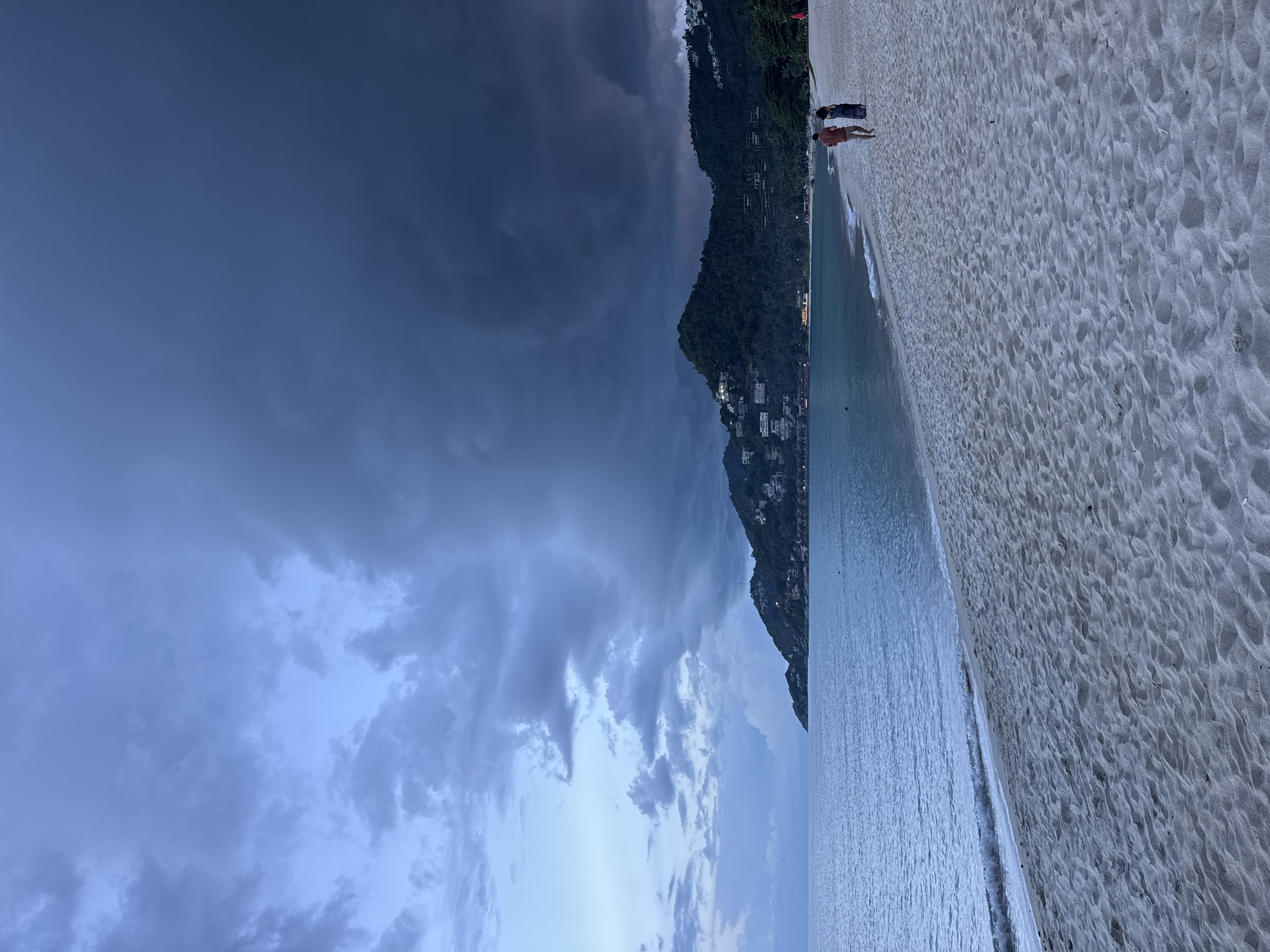 The image depicts a serene beach scene in Baan Chaweng Noi, Thailand, under a dramatic, overcast sky. The foreground is dominated by a wide expanse of fine, light-colored sand, marked with numerous small ripples and footprints, suggesting recent activity. Along the edge of the sand, the gentle waves of the ocean create a soft, white foam as they recede.
To the left of the sand, the tranquil turquoise water stretches out, its surface slightly rippled. Further to the left, a lush green mountainside rises steeply, dotted with numerous white buildings, indicating a populated coastal area or resort. The sky above the mountain is a deep, ominous gray, filled with heavy, swirling clouds that suggest an approaching storm or the aftermath of one. Towards the right side of the image, the sky transitions to a lighter, softer blue, hinting at a break in the weather or the approaching twilight.
Two individuals are visible on the sand, standing near the water's edge. One person is wearing a reddish-orange shirt and shorts, while the other is wearing dark clothing. Their posture suggests a relaxed, perhaps contemplative mood as they gaze out at the sea. The overall atmosphere is one of natural beauty contrasted with impending weather, evoking a sense of calm before a storm. The time of day appears to be late afternoon or early evening, given the quality of the light and the cloud formations. No visible text is present in the image.