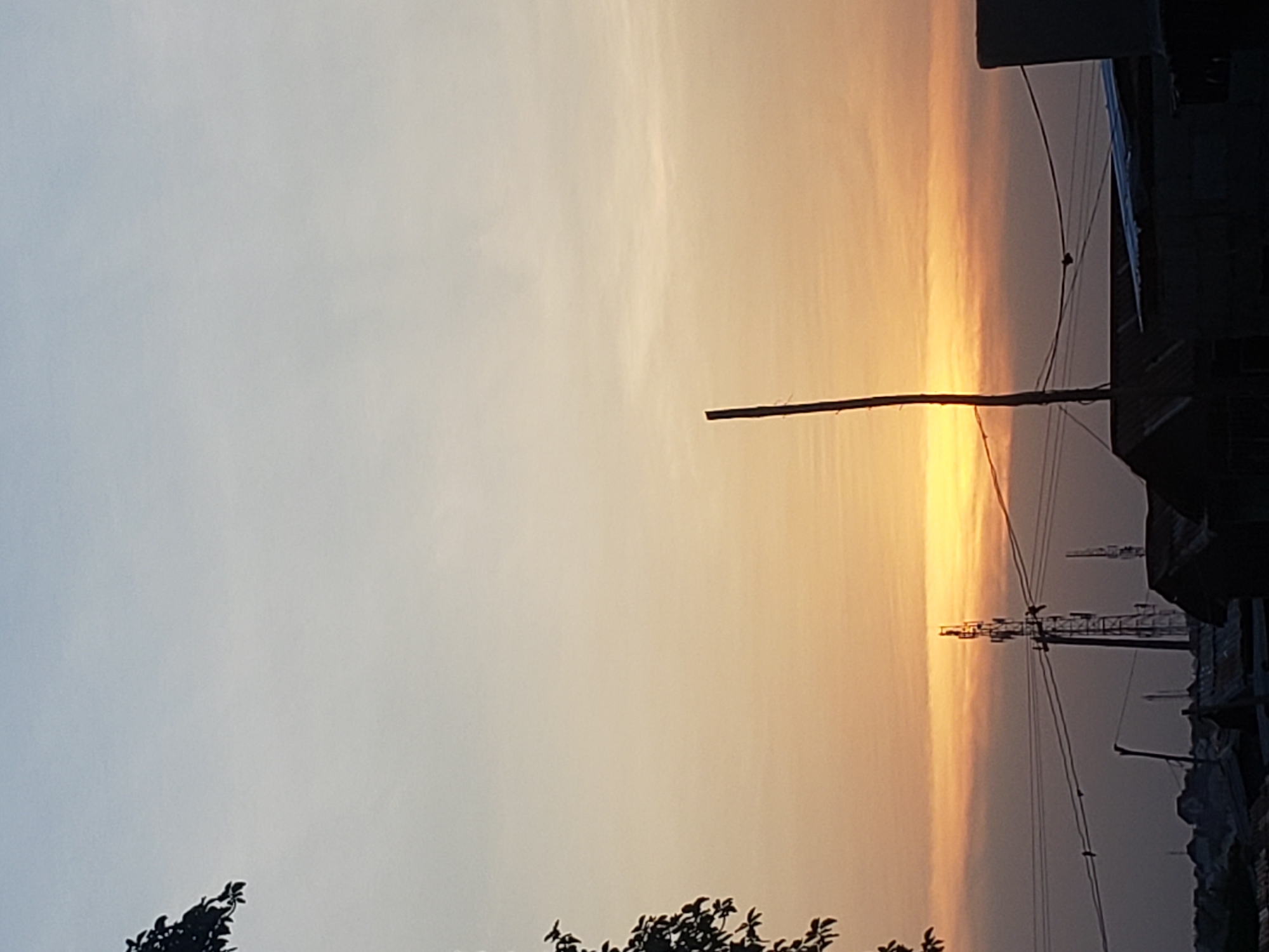 This panoramic image captures a vibrant sunset or sunrise over a populated area in Adankolo, Nigeria, characterized by warm hues and stark silhouettes. The sky dominates the frame, showcasing a beautiful gradient from a soft, pale blue-grey at the top to brilliant yellows, oranges, and deeper reds along the horizon line. Wispy, indistinct clouds are faintly visible, illuminated by the low-lying sun.
In the mid-ground, a tall, slender utility pole stands upright, appearing as a dark silhouette against the glowing sky. Multiple thin electrical wires extend horizontally from this pole, crisscrossing the frame. Further along the wires and in the distance, another, larger electricity pylon or communication tower is visible, also in silhouette.
On the far right side of the image, a cluster of buildings or structures are rendered in dark silhouette, suggesting an urban or semi-urban environment. Their varied rooflines and angular shapes create a stark contrast against the bright sky. Silhouetted tree branches and foliage are visible in the bottom corners, adding to the foreground elements.
The scene conveys a sense of calm and natural beauty often associated with the transition periods of day and night. There is no visible text in the image.
