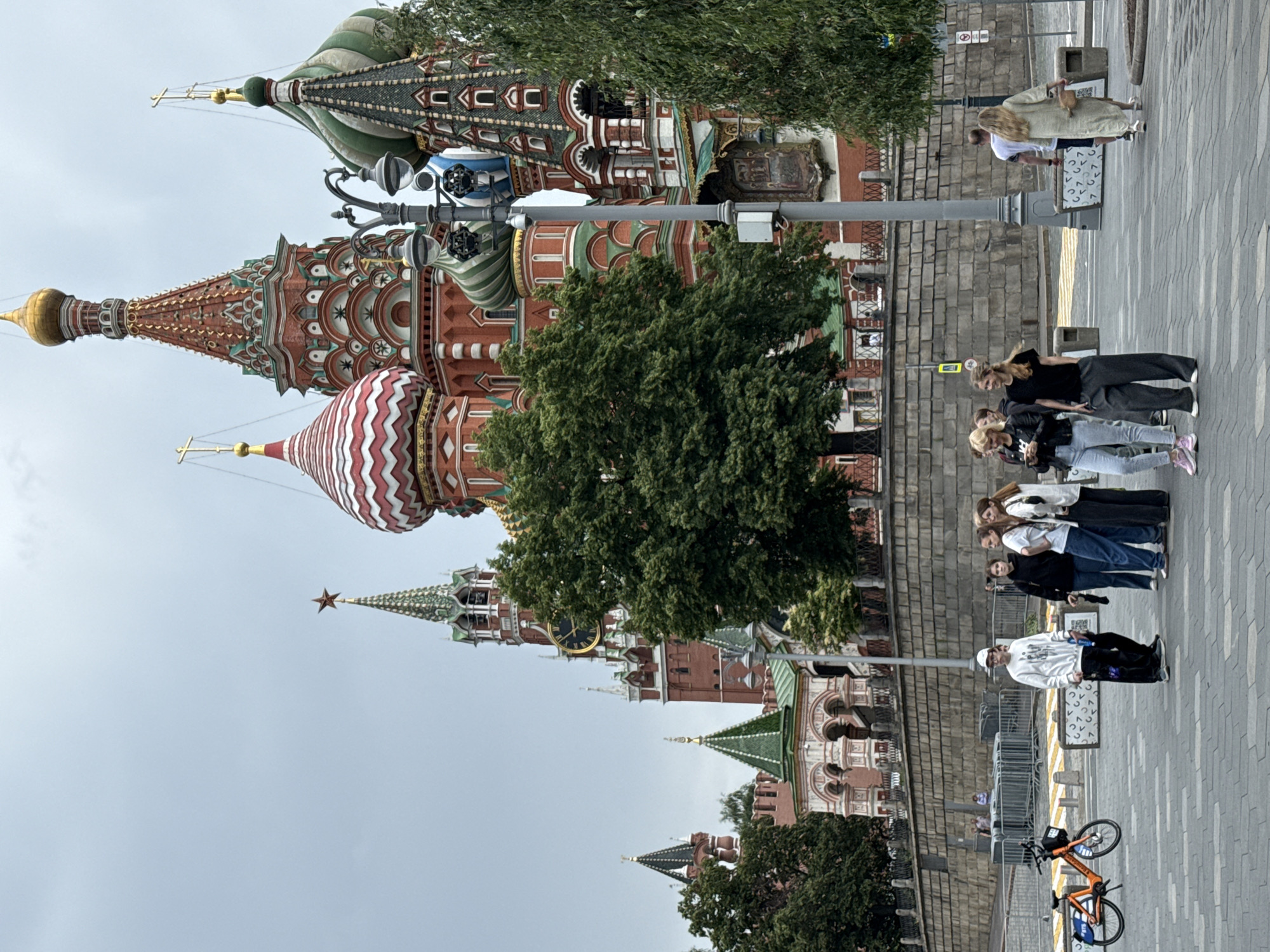 The image shows a group of tourists standing in Red Square, Moscow, Russia, in front of Saint Basil's Cathedral. The sky is overcast, and the architecture of the cathedral is prominent with its colorful onion domes and spires. Several people are visible, some looking towards the cathedral, others talking amongst themselves. In the foreground, a few individuals are walking or standing around. An orange bicycle with "Яндекс" branding is parked to the left. The ground is paved with cobblestones. The scene captures a typical tourist experience in a historic landmark.