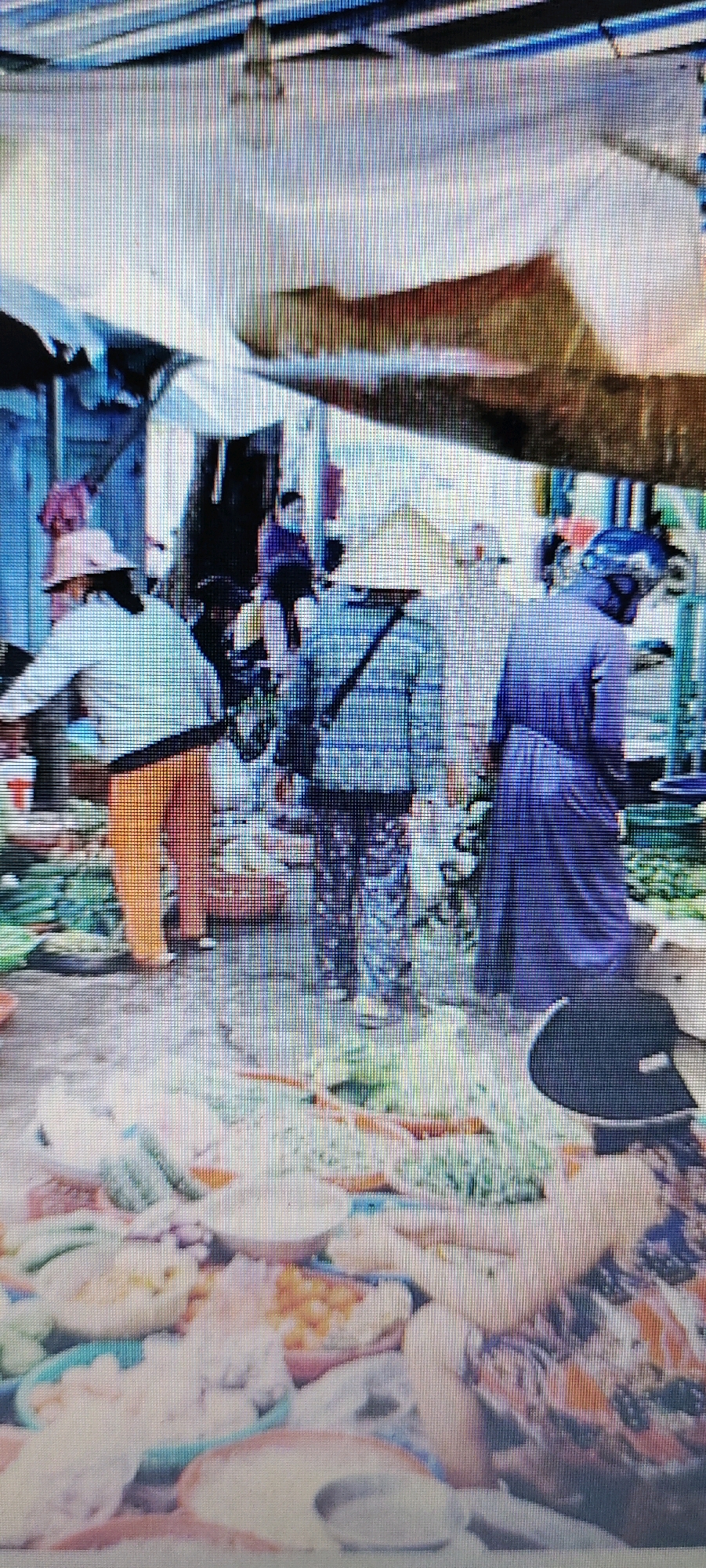 The image depicts a bustling outdoor market scene in Hendrosari, Indonesia. Several individuals, mostly women, are engaged in activities related to the market. One person on the left wears a light-colored jacket and orange pants, with a hat that appears to be made of straw. Another individual in the center wears a blue and white checkered shirt and patterned pants, also wearing a hat. To the right, a person in a dark blue, long-sleeved garment is visible. The foreground is filled with various food items arranged in bowls and on the ground, suggesting a food market. The background shows stalls and structures covered with tarps or awnings, providing shade. The overall impression is one of a busy commercial area with people actively participating in a transaction or preparing goods.