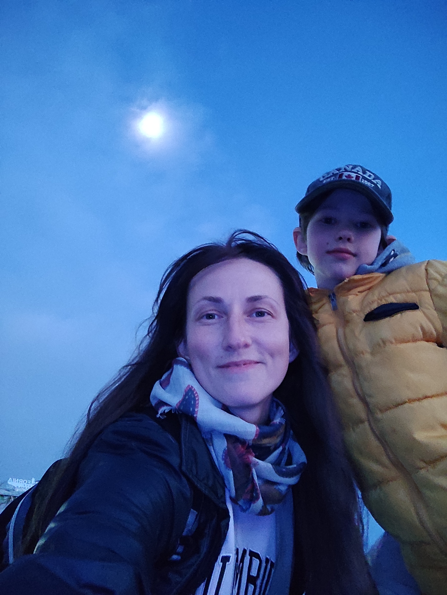A selfie-style photo captures a woman and a younger person, likely a child, against a backdrop of a blue sky and what appears to be a bright moon or light source. The woman is in the foreground, wearing a dark jacket, a scarf with a patterned design, and a light-colored shirt with visible writing. Her hair is long and dark. The younger person is positioned behind and to the right of the woman, wearing a yellow jacket and a baseball cap with a "CANADA" logo. The sky above is a clear blue with a single bright light source, possibly the moon or a strong artificial light. The location is described as Sochi, Russia, though specific landmarks or contextual elements that confirm this are not clearly visible in the image.