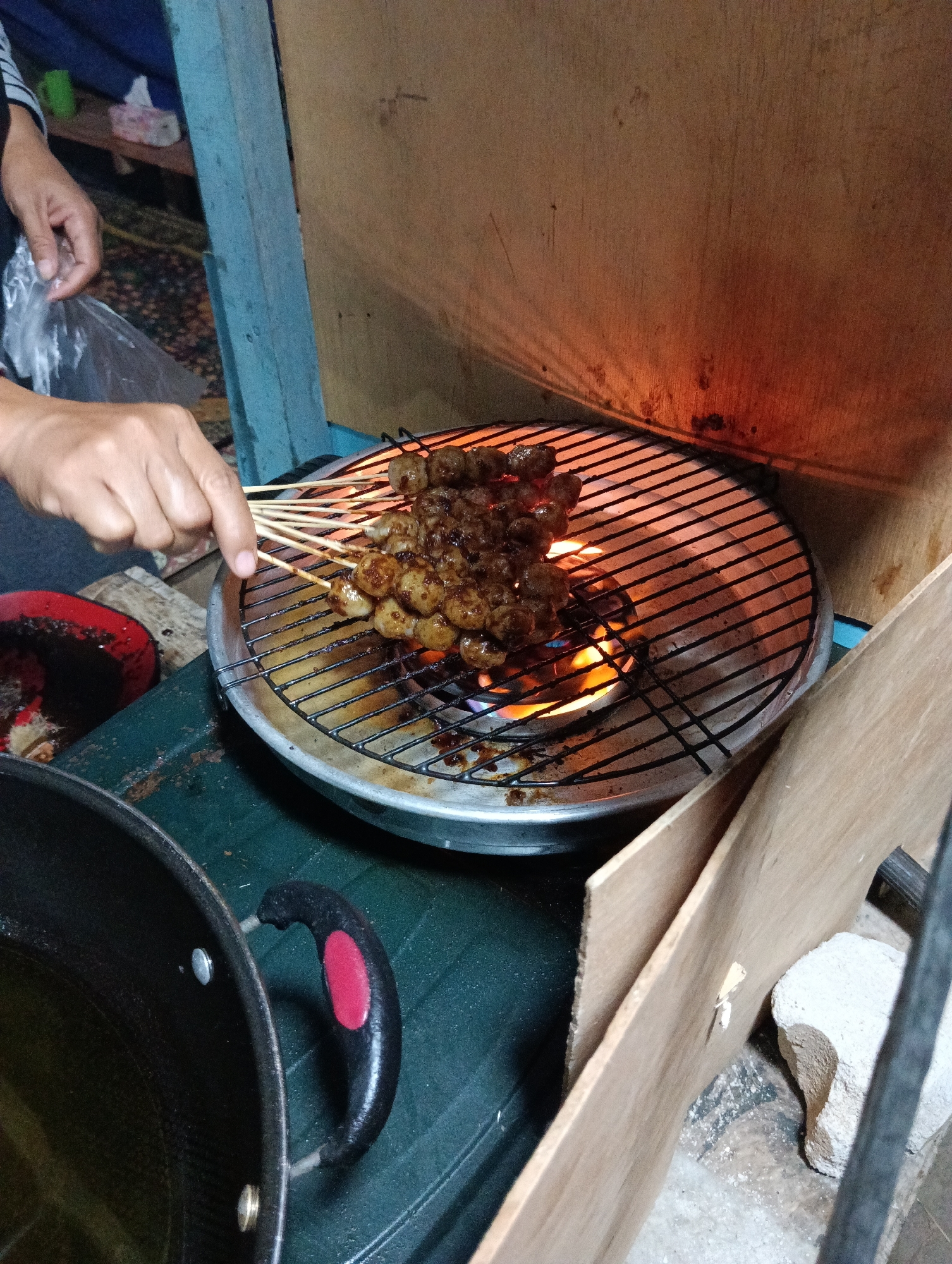 In Pematang Jaya, Indonesia, a person's hands are visible, holding plastic wrap while a charcoal grill cooks skewers of meatballs over an open flame. The grill is circular and sits on a green surface, possibly a table or counter. The meatballs are glazed and appear to be seasoned. In the foreground, a large black pot with a red and black handle is visible, containing what looks like cooking oil. The background is somewhat dark and cluttered, with hints of blue and brown structures, suggesting an outdoor or semi-outdoor food stall setting. The lighting is dim, with the glow of the grill providing the primary illumination.