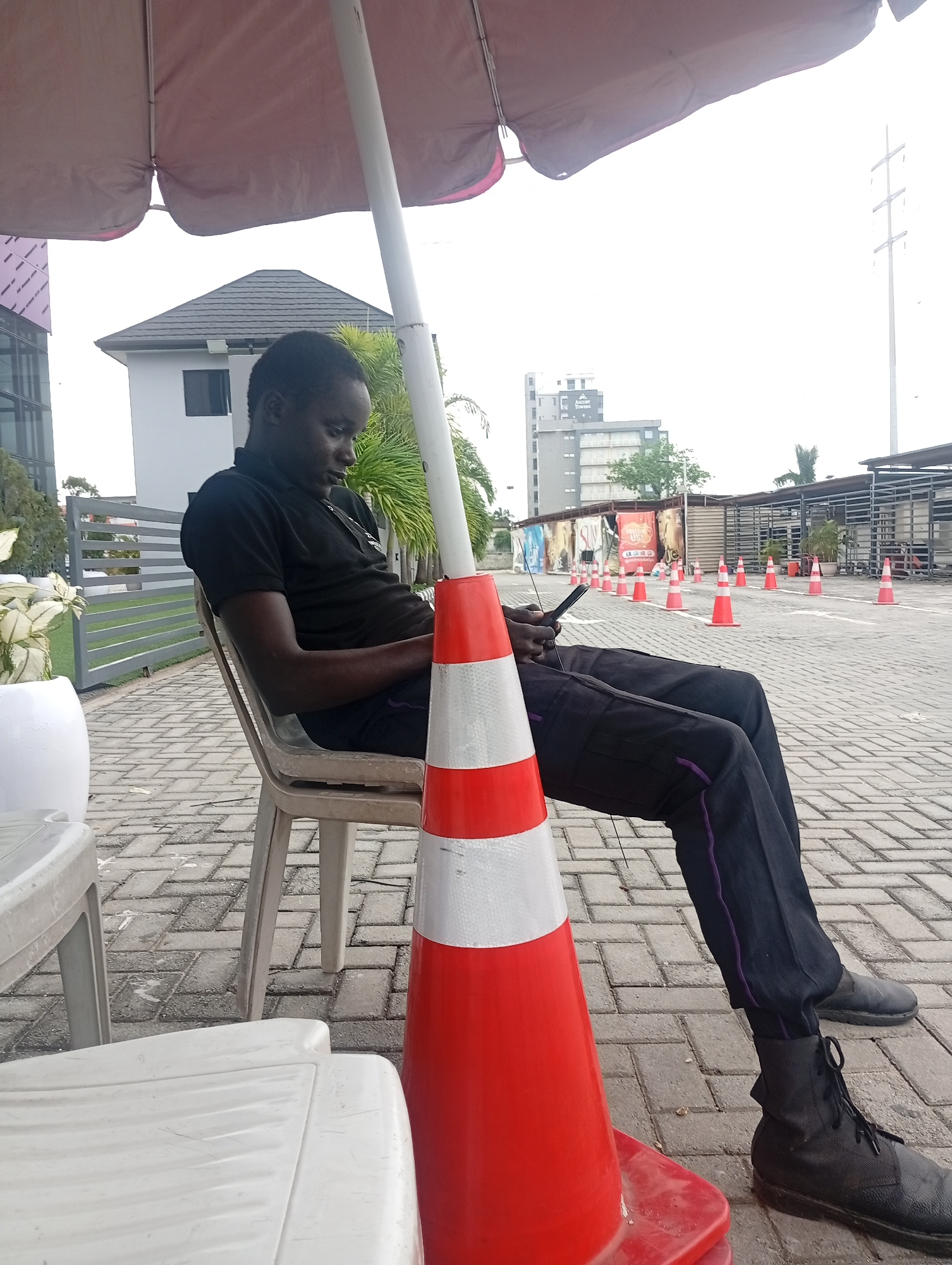 A person wearing a black polo shirt and dark pants with a purple stripe is sitting on a plastic chair under a large umbrella. They are holding a mobile phone in their hands, looking down at it. A large orange traffic cone with white reflective bands is positioned in front of the chair. The ground is paved with grey interlocking bricks. In the background, there are several other orange traffic cones lined up, along with some covered structures and buildings, including a tall grey building with "Ascot Towers" written on it. A tall electricity pylon is visible in the distance against a bright sky. White plastic tables and chairs are partially visible in the foreground on the left. The scene appears to be an outdoor area, possibly a parking lot or a waiting area in Ilado, Nigeria.
